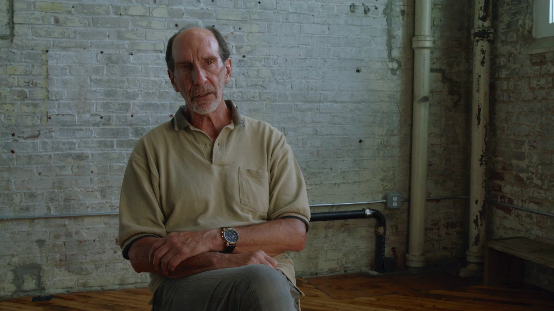 An older man with short hair and a beard, wearing a beige polo shirt with dark trim, sitting on a wooden floor against a white brick wall. He has his arms crossed and wears a wristwatch. There are pipes and an electrical outlet visible on the right side of the wall.