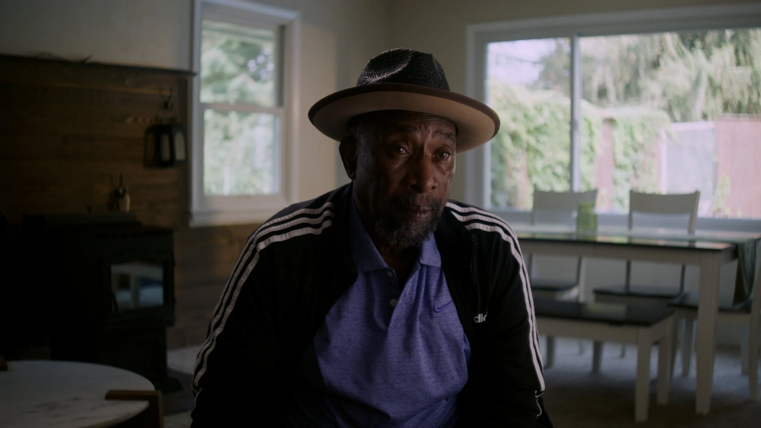 An older man with a beard and moustache wearing a wide-brimmed hat, jacket, and polo shirt, sitting indoors with large windows showing trees outside.