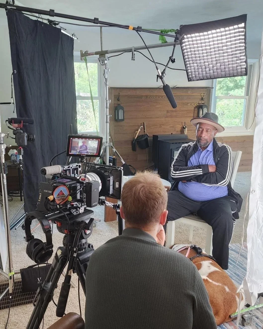 Man speaks during an interview or film shoot, seated by a window, with filming equipment, crew member, and camera setup in the background.