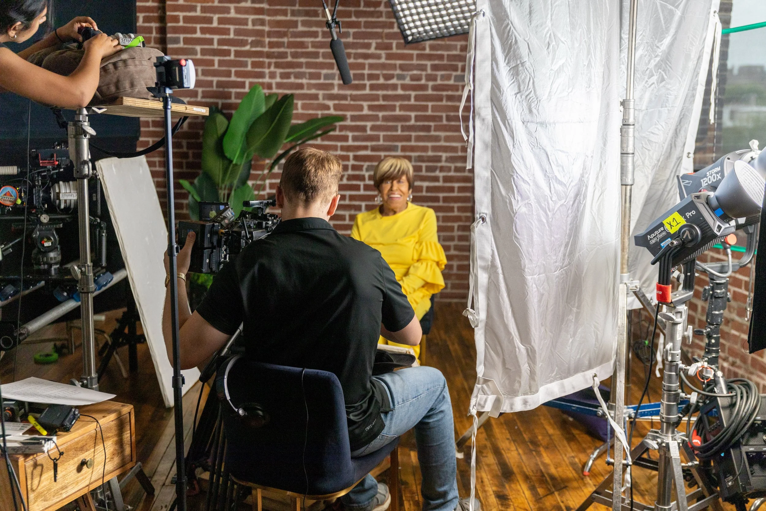Film production set with crew filming woman wearing yellow dress against brick wall, surrounded by cameras, lights, and equipment.