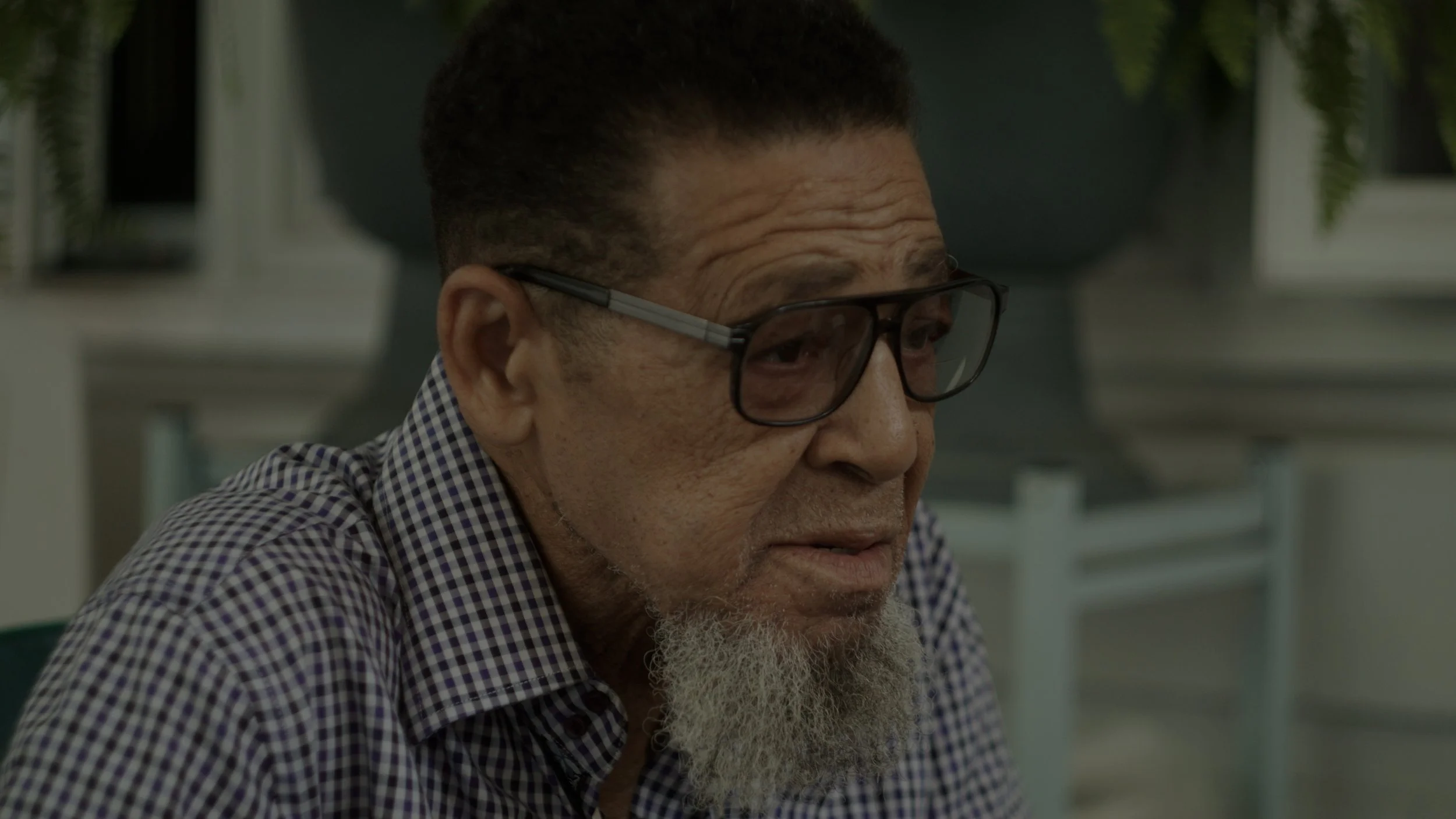 Close-up of an elderly man with a beard and glasses wearing a checkered shirt, sitting indoors.