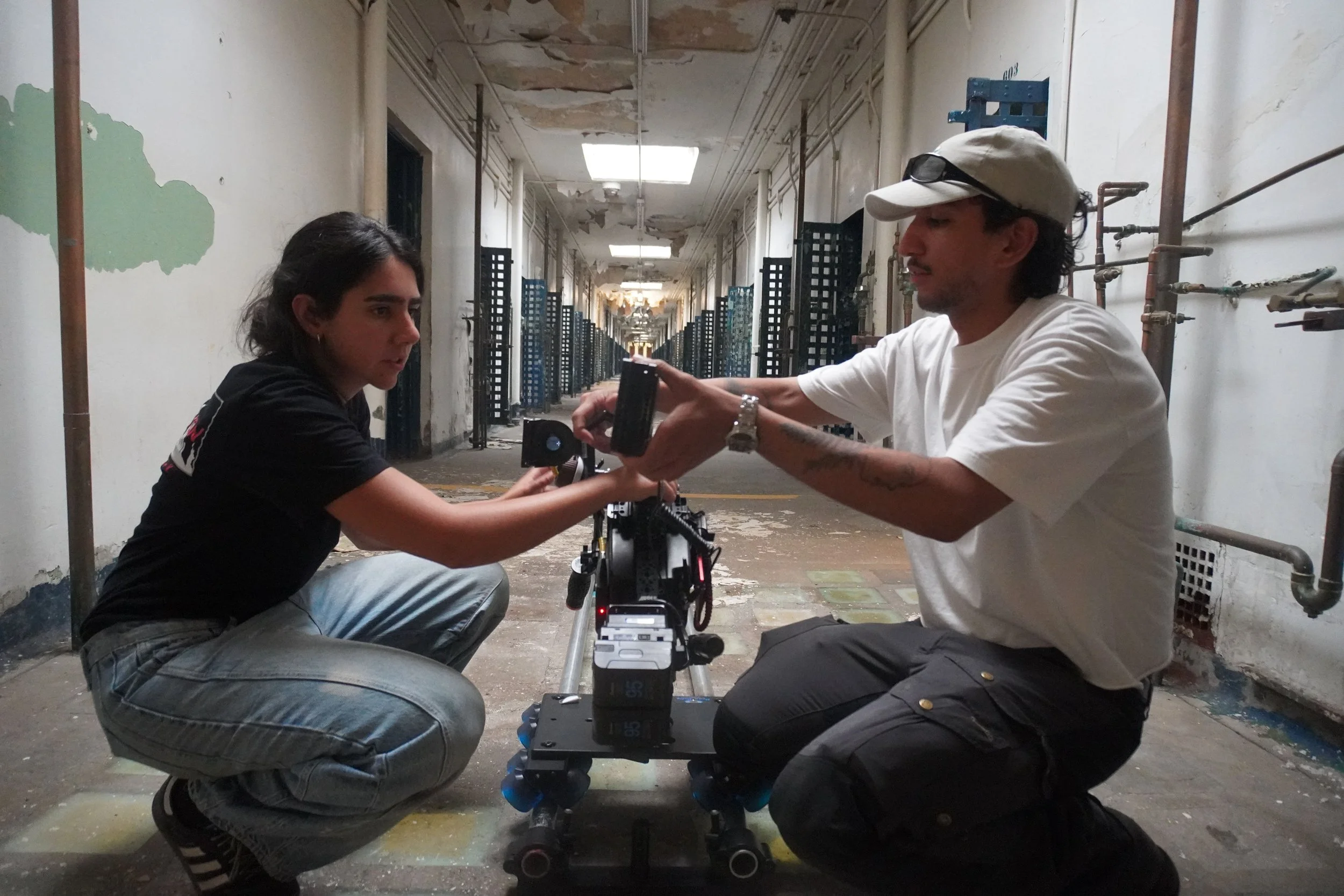 Two filmmakers setting up a camera on a tripod in a dilapidated hallway with peeling paint on the ceiling and walls, and exposed pipes.