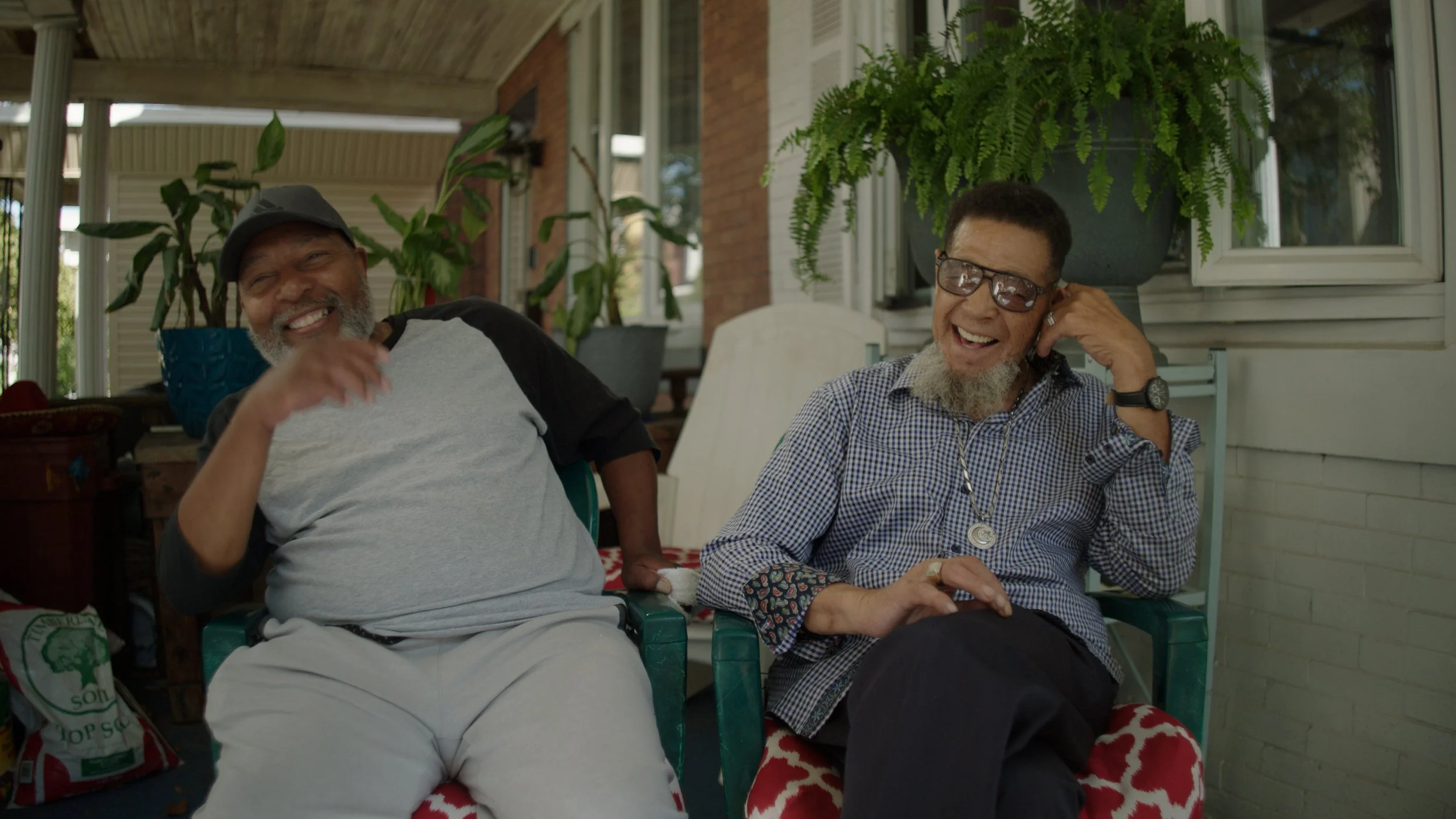 Two men sitting on porch chairs, smiling and laughing. One man wears a gray and black T-shirt and gray cap, the other man wears glasses and a checkered shirt. Potted plants and brick house wall are in background.