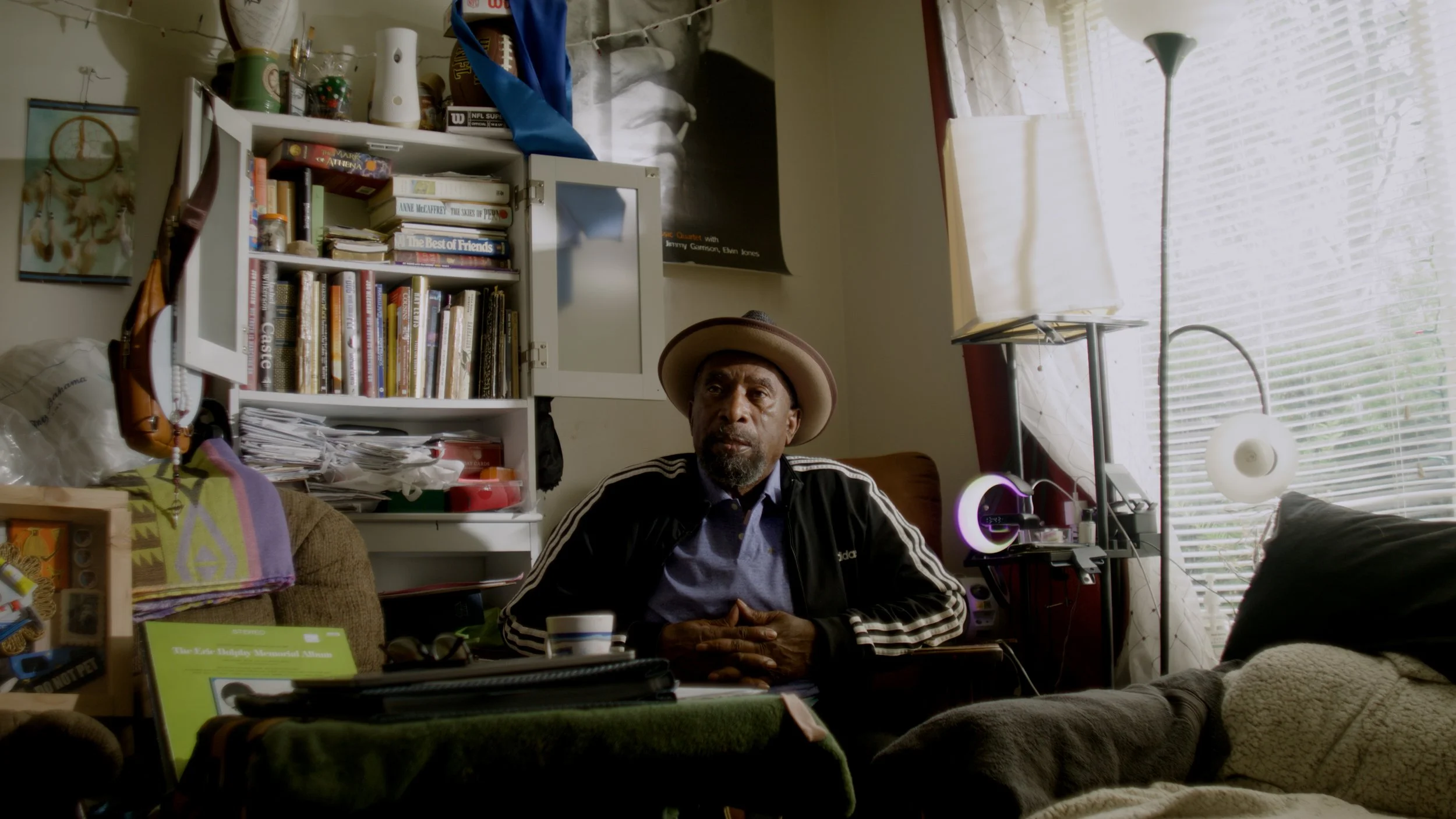 A man wearing a hat and a black Adidas jacket sitting at a cluttered desk in a room with bookshelves, large windows with blinds, and various items on the shelves and table.