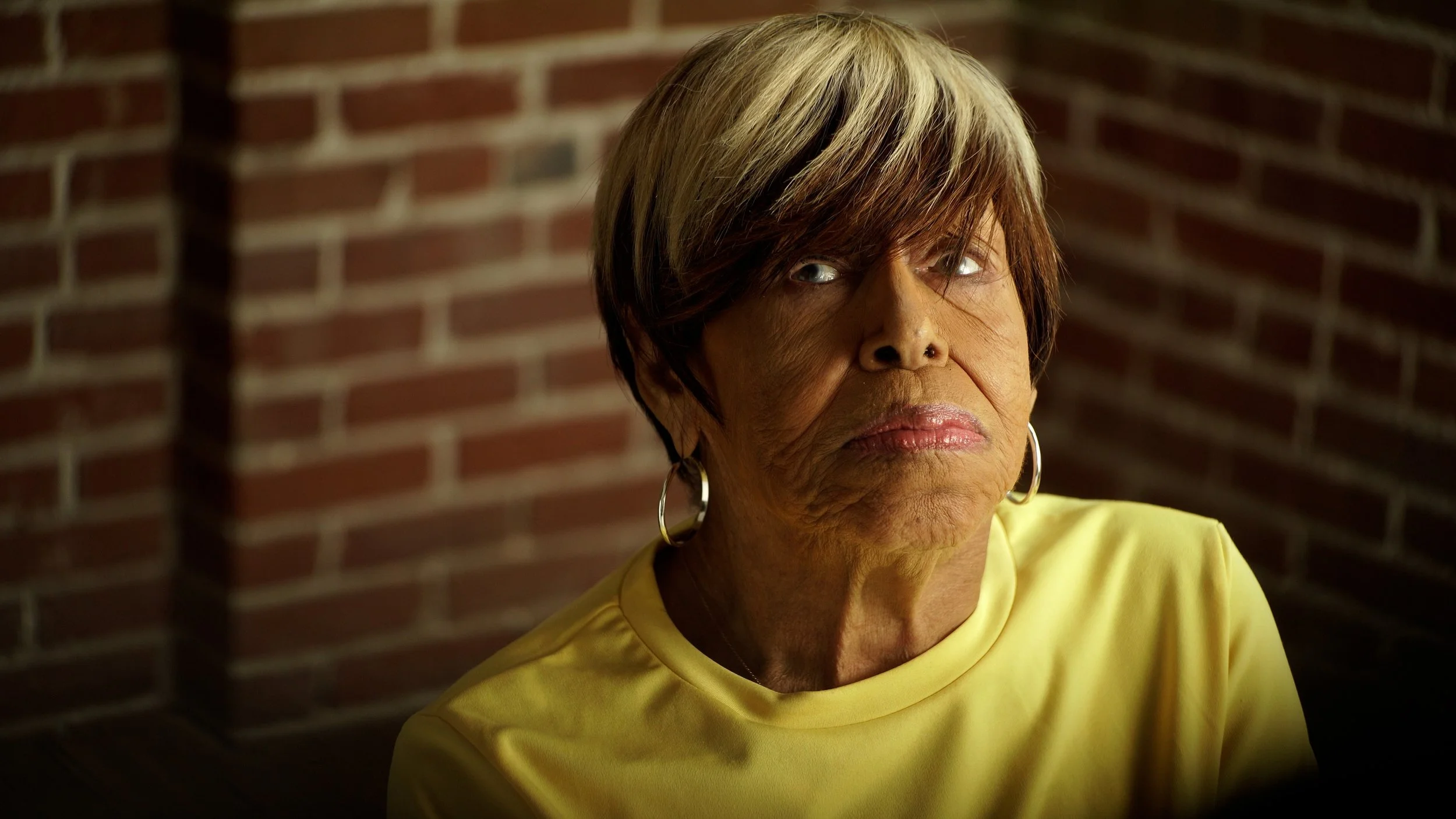 An older woman with short, styled hair and hoop earrings wearing a yellow shirt and standing in front of a brick wall.