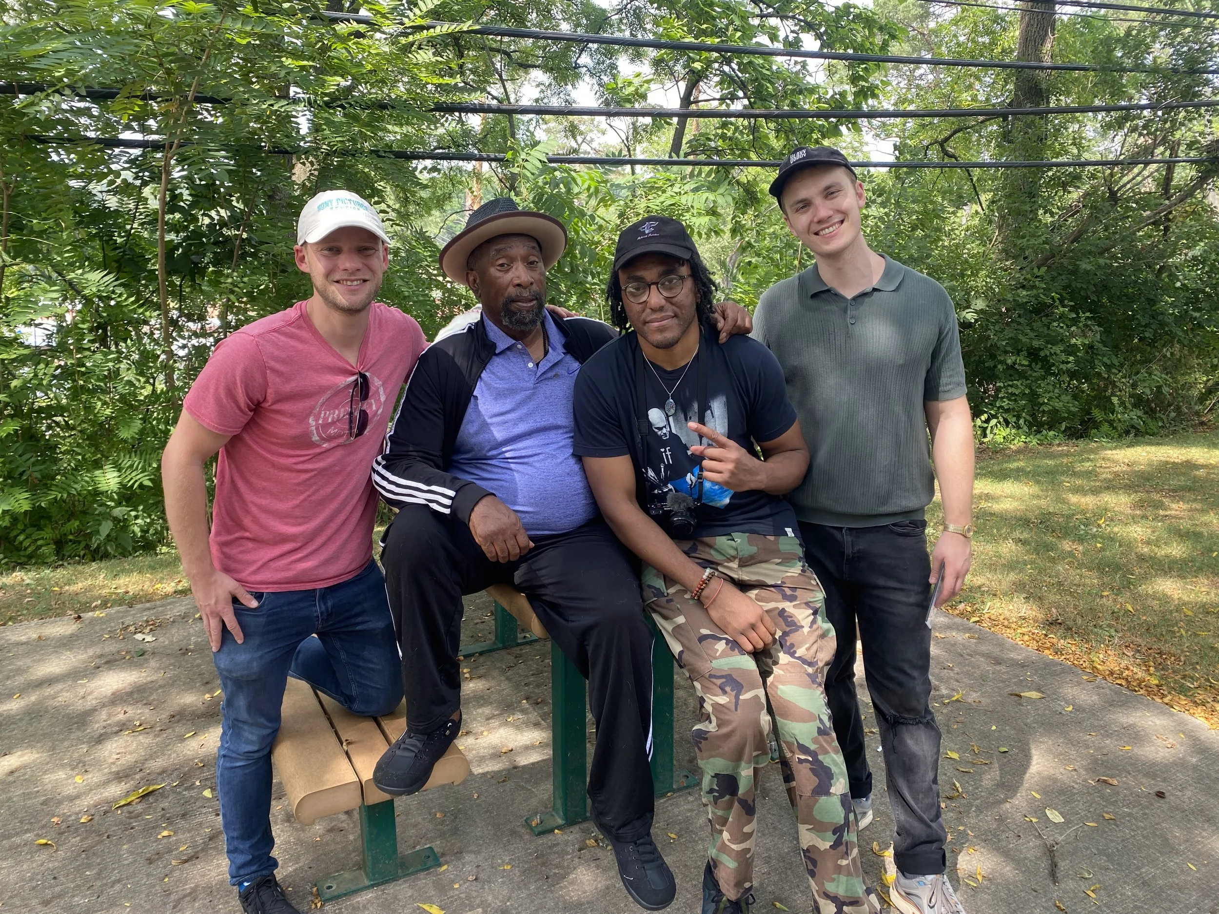 Four men standing together outdoors, smiling. Two are wearing hats. One has glasses. They are in a green, leafy park with trees and sunlight.