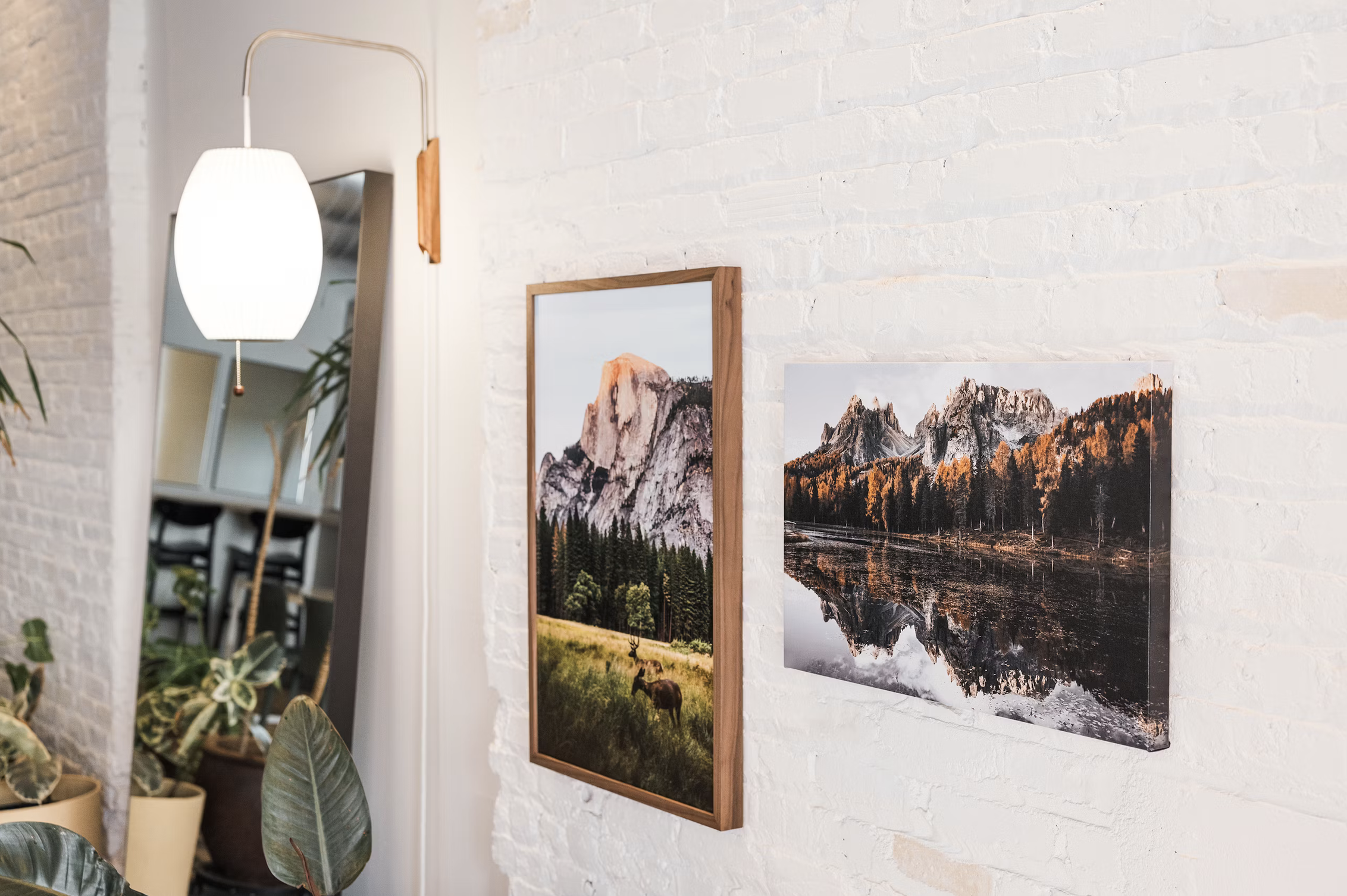 Interior of a room with white brick walls and framed landscape photographs of mountains and lakes hanging on the wall. A lamp with a white shade is turned on, illuminating the space. Several houseplants are visible in the foreground.