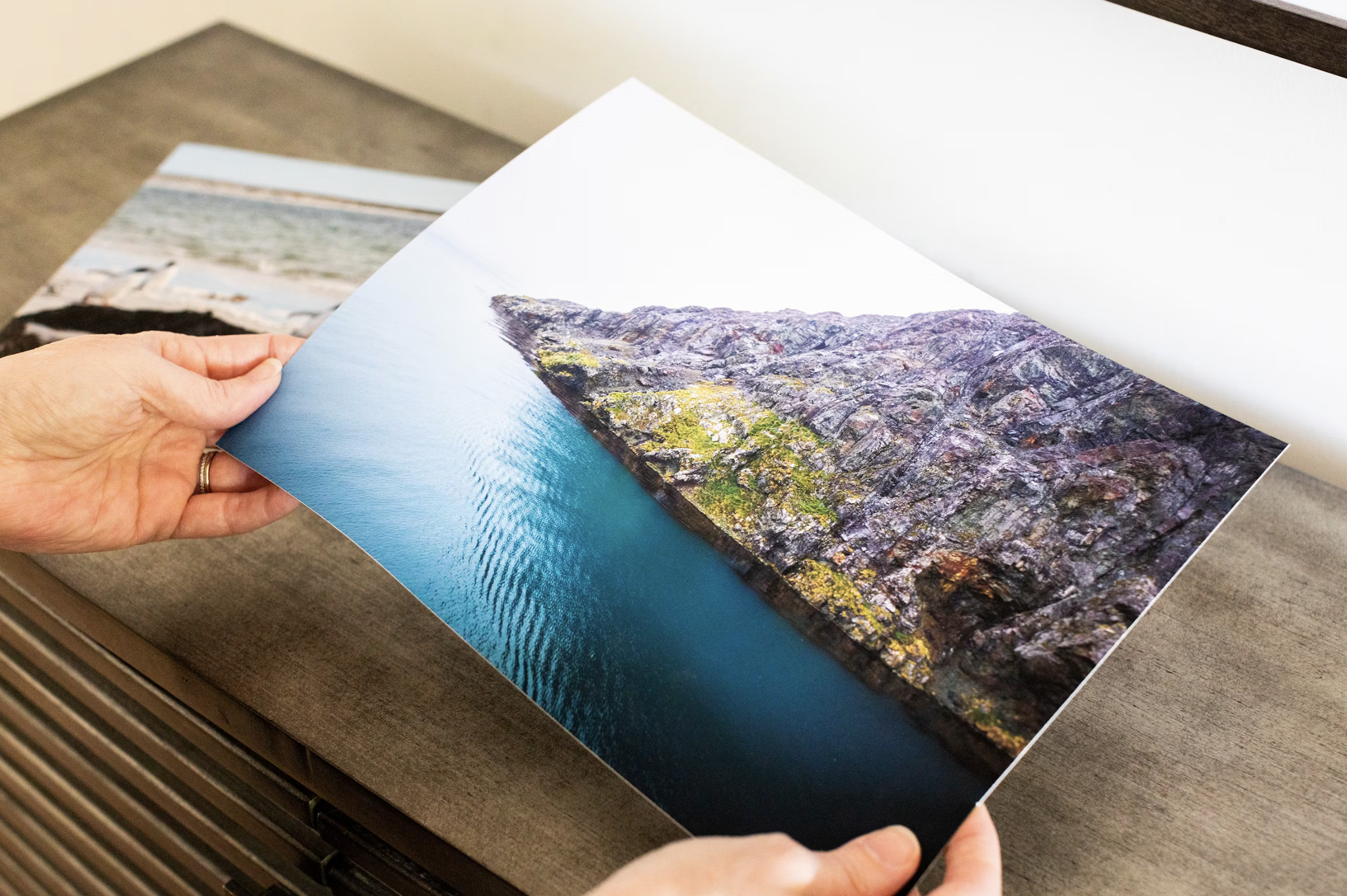 A person holding a printed photograph of a mountain and lake landscape, with the photo partially folded.