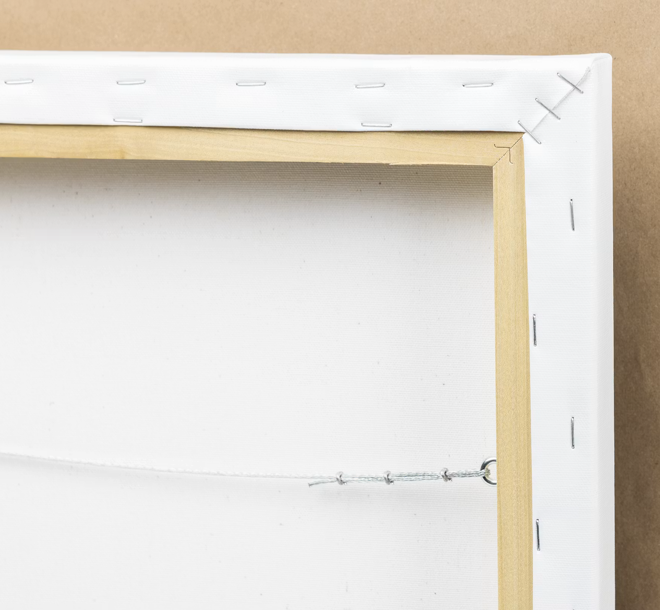 Close-up of the back of a stretched canvas, showing wooden frame, white fabric, staples, and wire for hanging, with brown paper background.