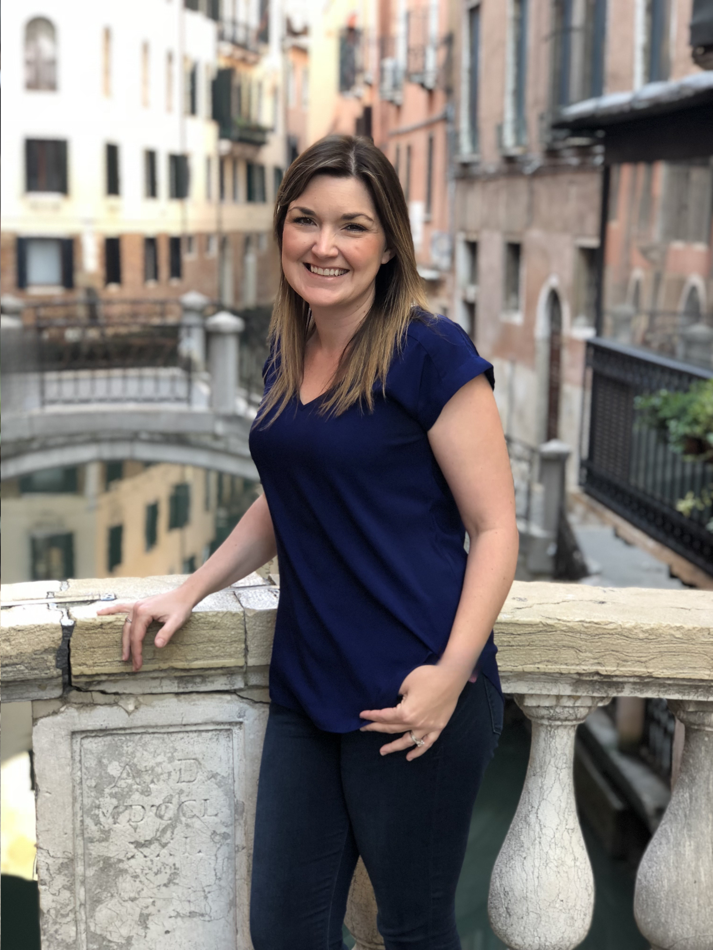 A smiling woman with shoulder-length brown hair wearing a blue top and dark jeans, standing on a balcony with a stone railing, overlooking an urban canal lined with old brick buildings.