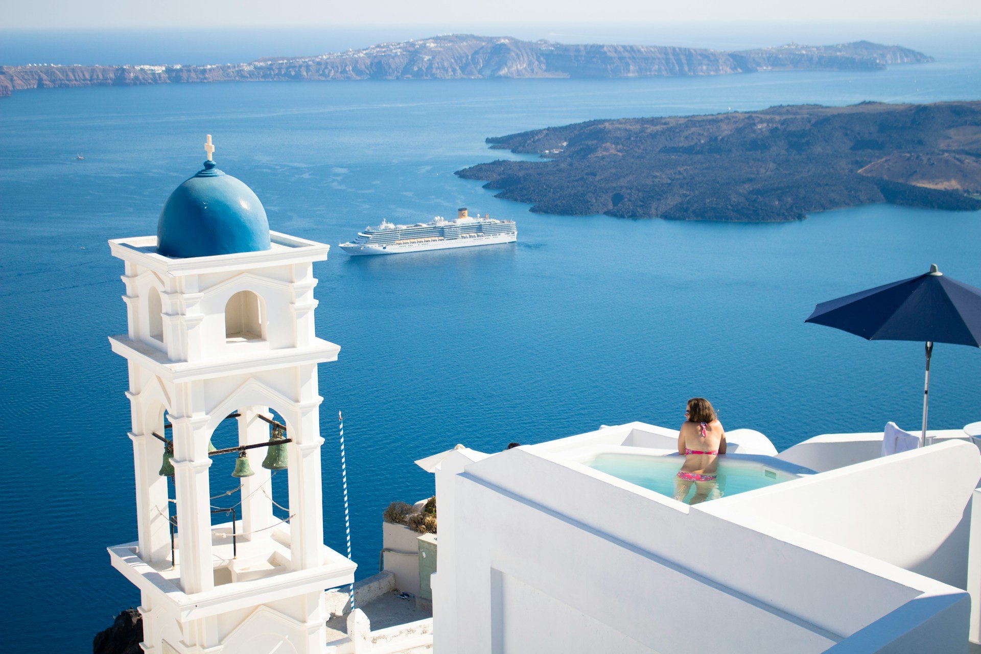 A woman relaxing in a small pool on a white rooftop overlooking the blue Aegean Sea in Santorini, Greece, with the iconic white buildings, church bell tower, and a cruise ship in the distance.