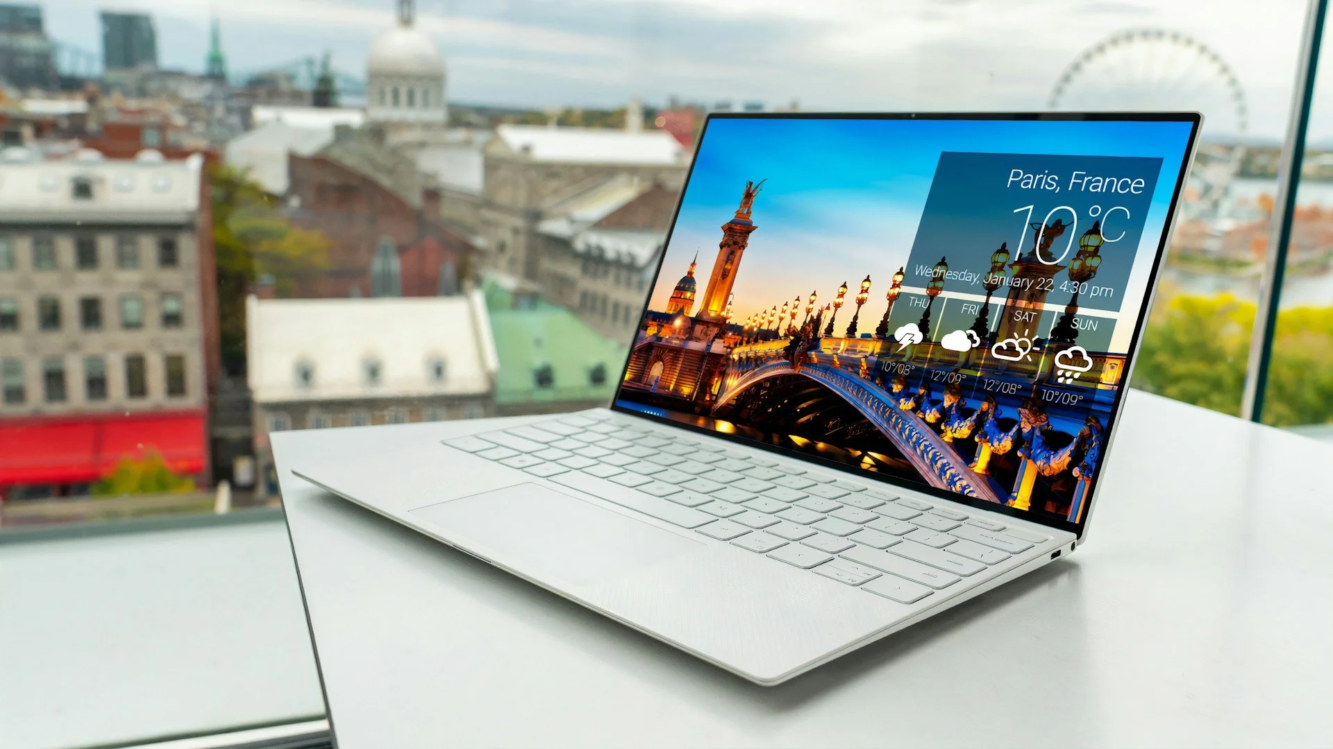 A white laptop on a white table in front of large windows with a cityscape view. The laptop screen displays a weather widget for Paris, France, showing 10°C and a picture of a bridge and city lighting at sunset.