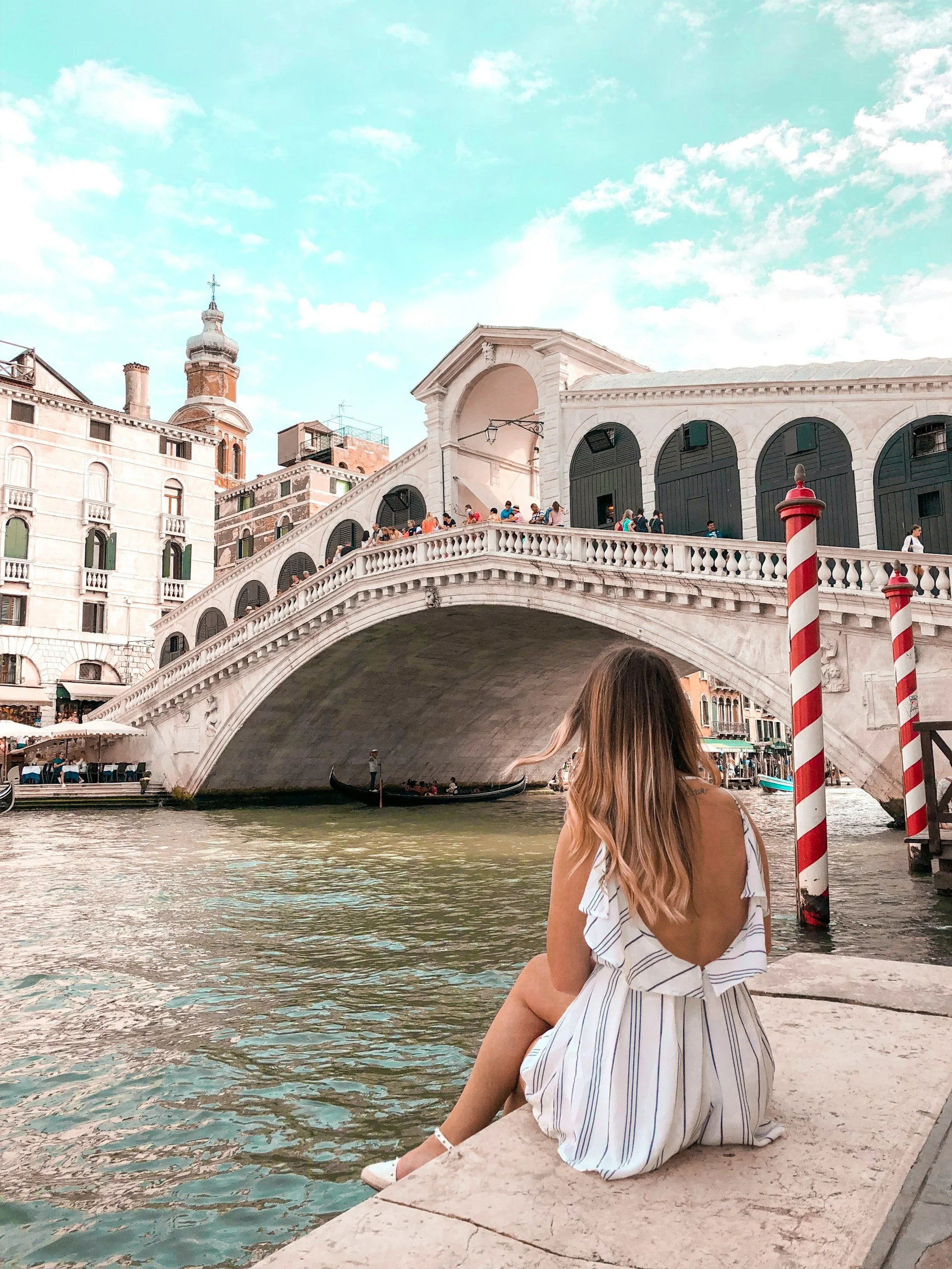 A woman with long hair wearing a striped dress is sitting on a stone ledge by a canal, with the Rialto Bridge in Venice, Italy, in the background. The bridge has a white stone design with arches and a staircase, and people are walking on it. The sky is partly cloudy with a light blue color.