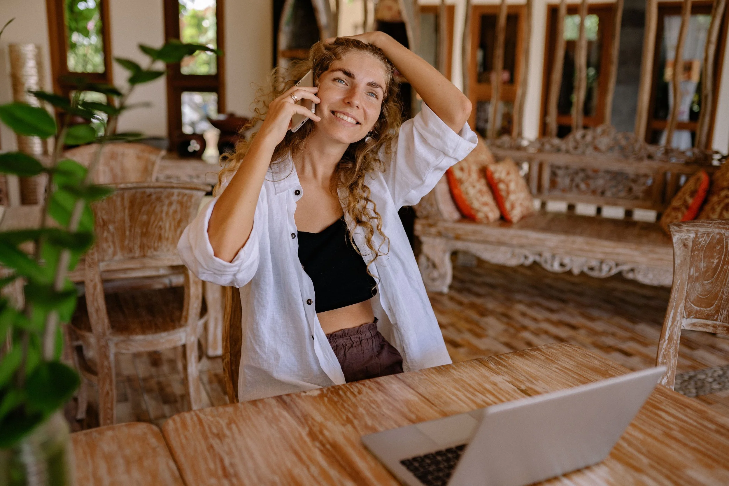 A woman smiling on a phone call at a wooden table in a resort setting, planning her travel itinerary.