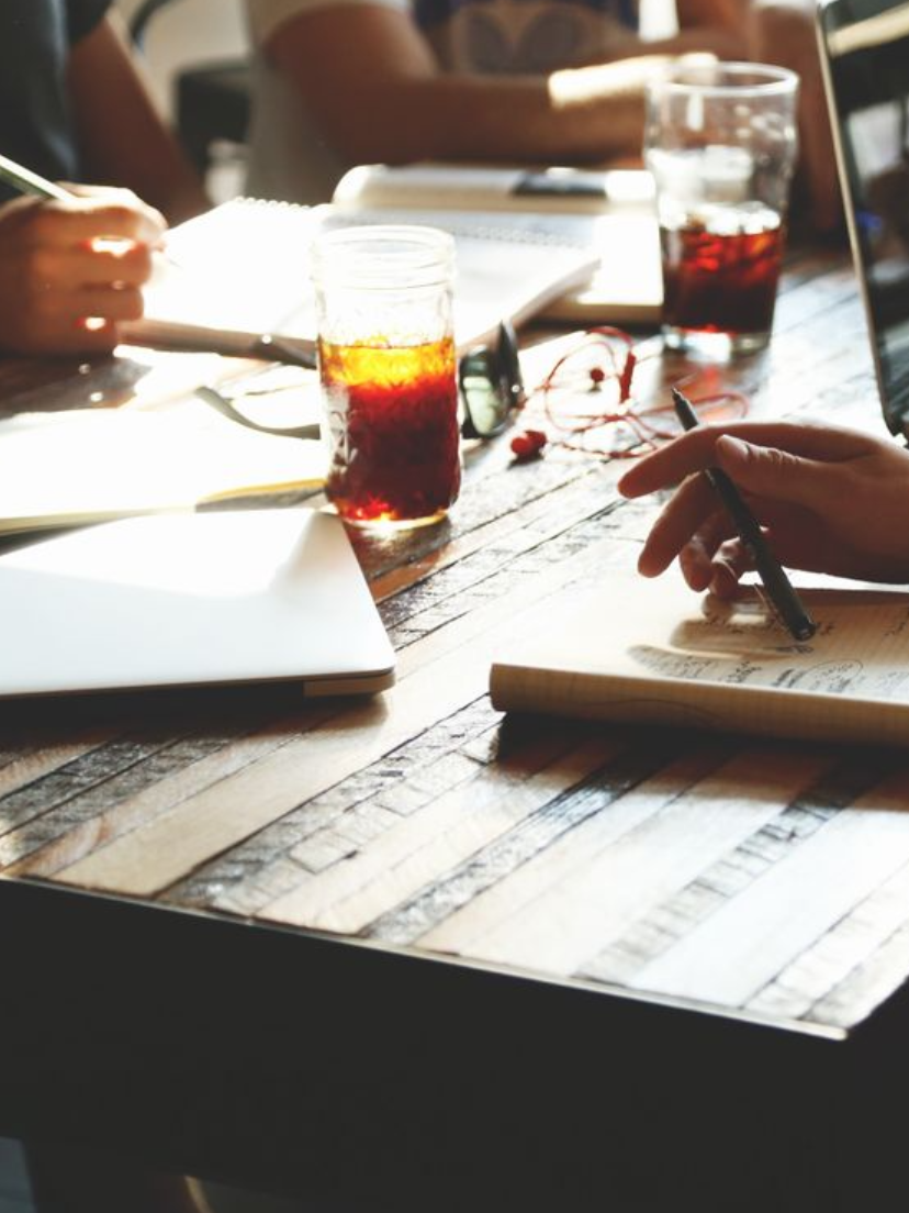People sitting at a wooden table with notebooks, pens, glasses of dark soda, and a laptop.