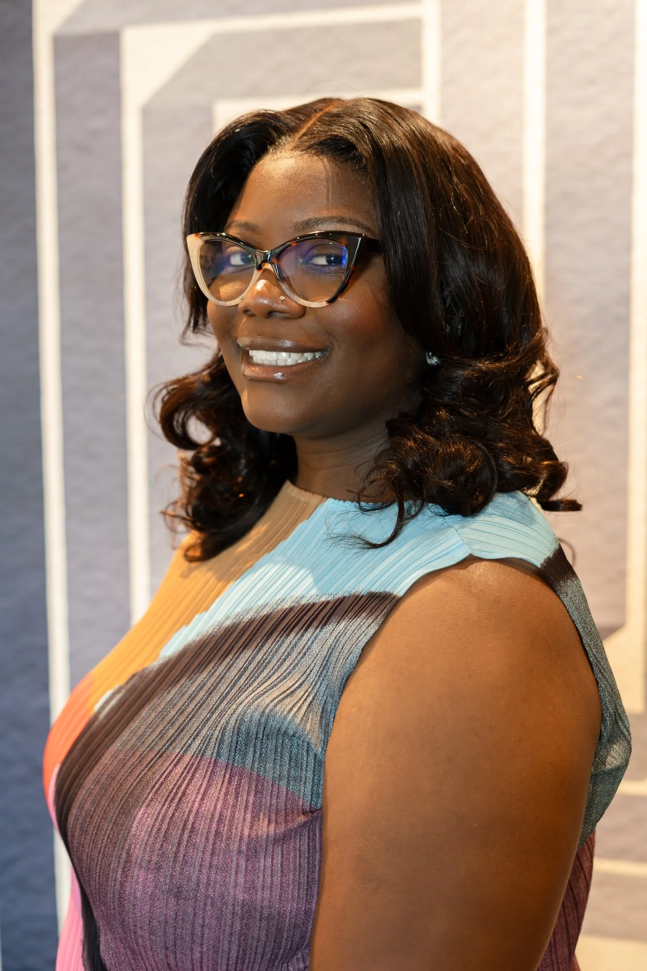 A woman with dark, wavy hair, wearing large tortoiseshell glasses and a sleeveless multicolored striped dress, standing indoors with a light gray wall and white vertical lines in the background.