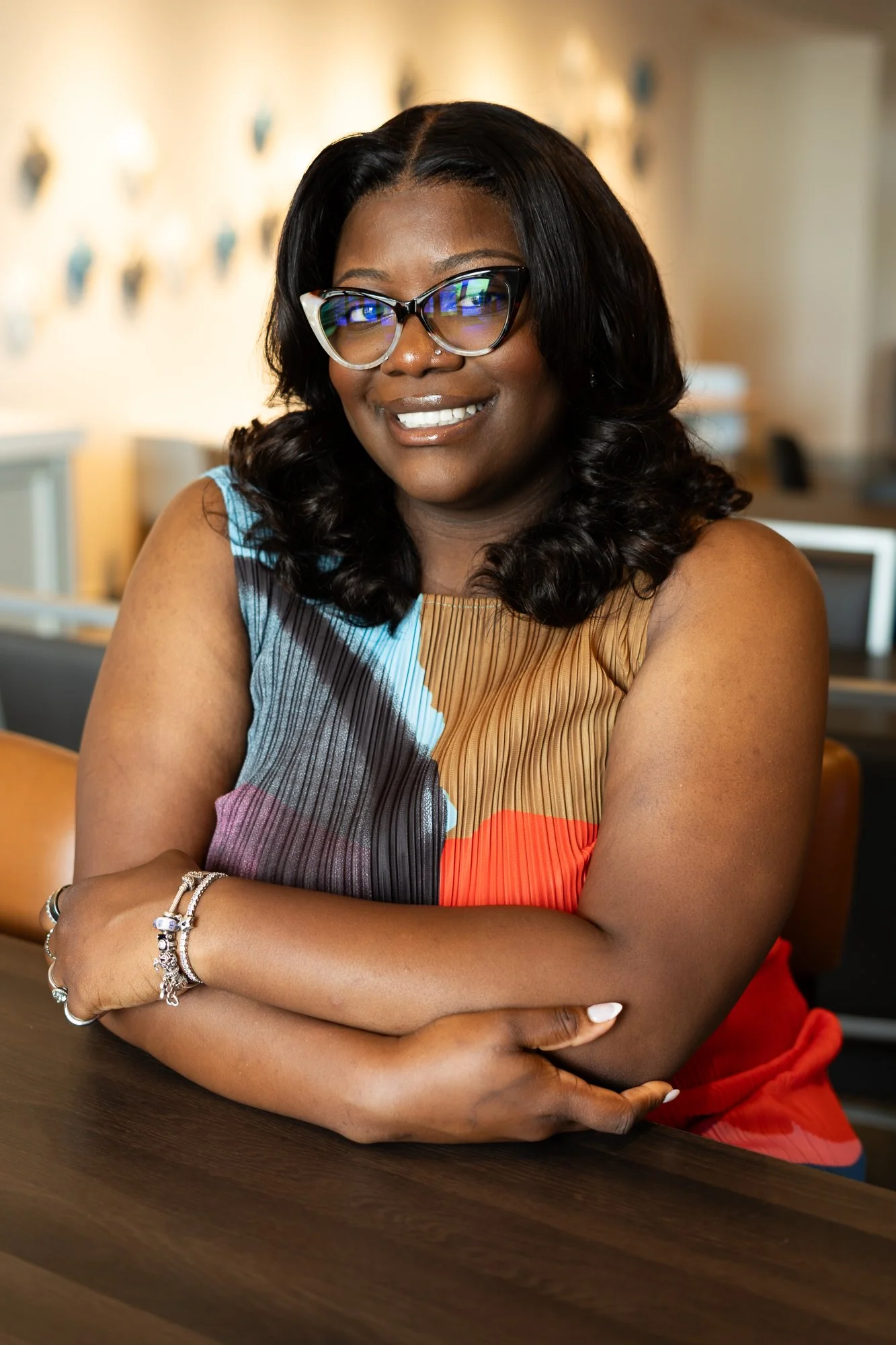 A woman with dark hair and glasses smiling with arms crossed, sitting at a wooden table inside a warmly lit room.