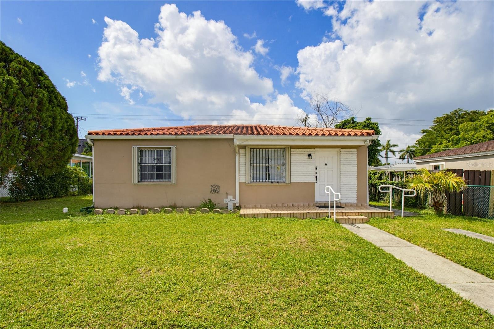 Front view of a small beige house with a red-tiled roof and white trim, surrounded by a green lawn and some trees, under a partly cloudy blue sky.