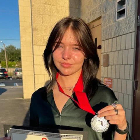 Young woman with shoulder-length brown hair holding a silver medal with a green button-up, standing outside a school.