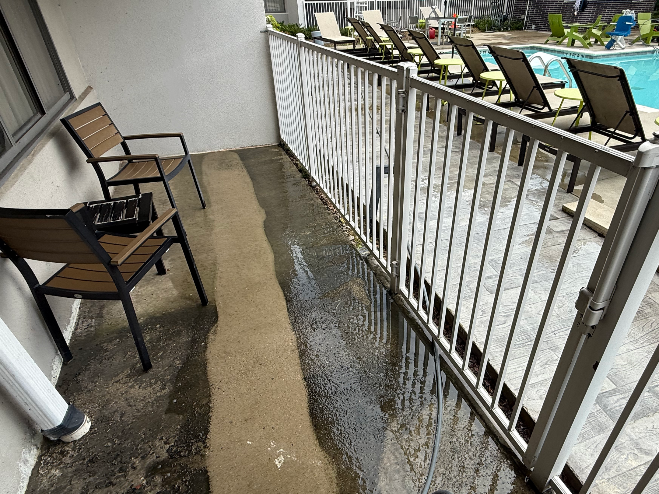 Balcony with outdoor chairs, a small table, and a pool in the background, after rain with wet floor.