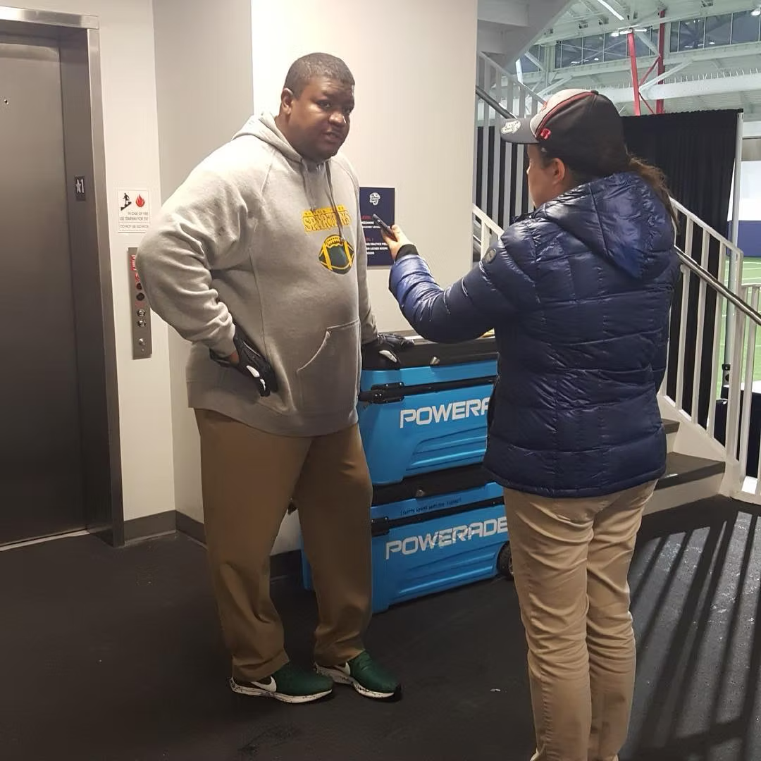 A man being interviewed by a woman at an indoor sports stadium. The man is wearing a gray hoodie and khaki pants, while the woman is dressed in a navy quilted jacket and beige pants. They are near luggage carts branded 'Powerade' and an elevator.