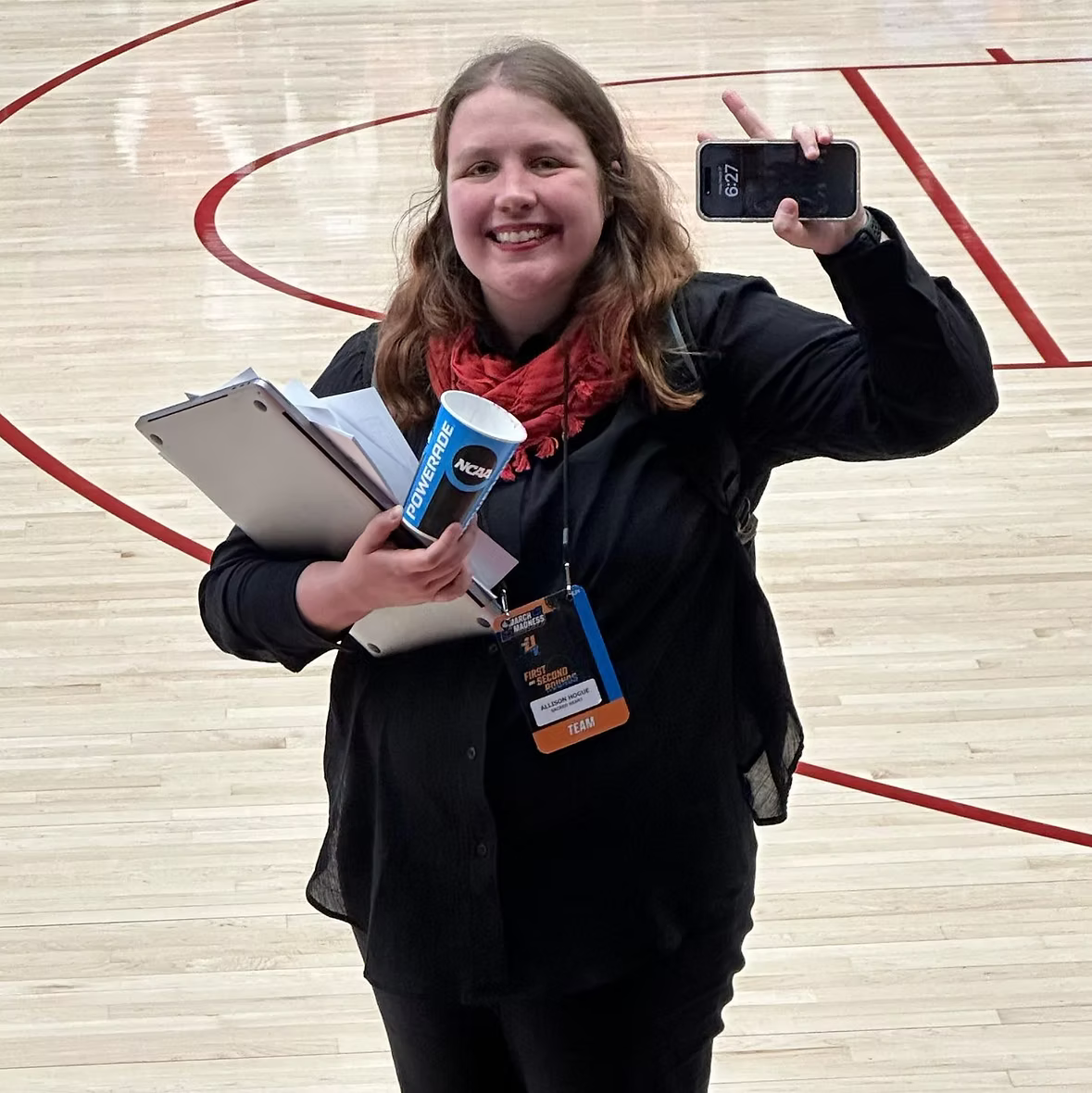 Smiling woman in black jacket with a red scarf taking a selfie on a basketball court, holding papers, a cup, and wearing event badge that says 'team'.