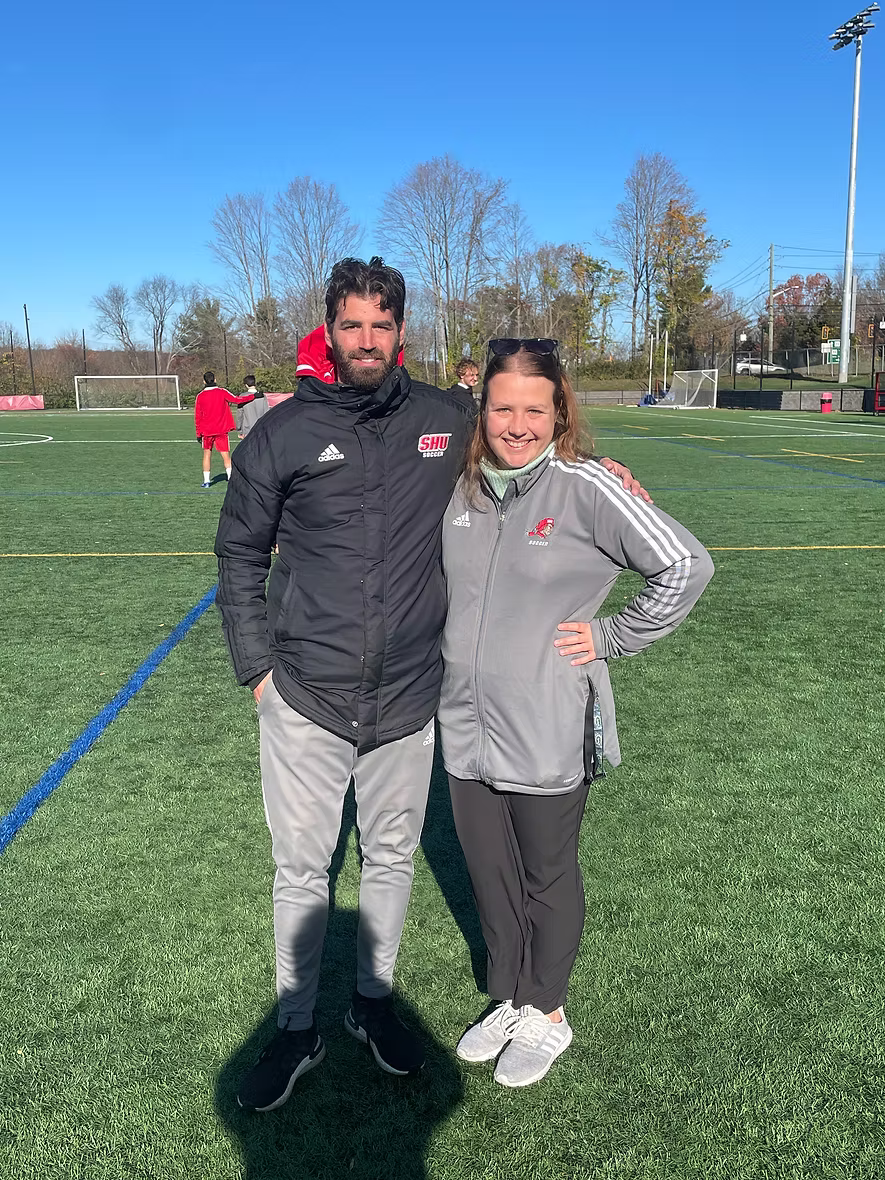 Two people, a man and a woman, standing on a soccer field on a sunny day. They are smiling, wearing sports jackets with the SHU logo, and have their arms around each other. In the background, there are other people on the field, trees, and goal posts.