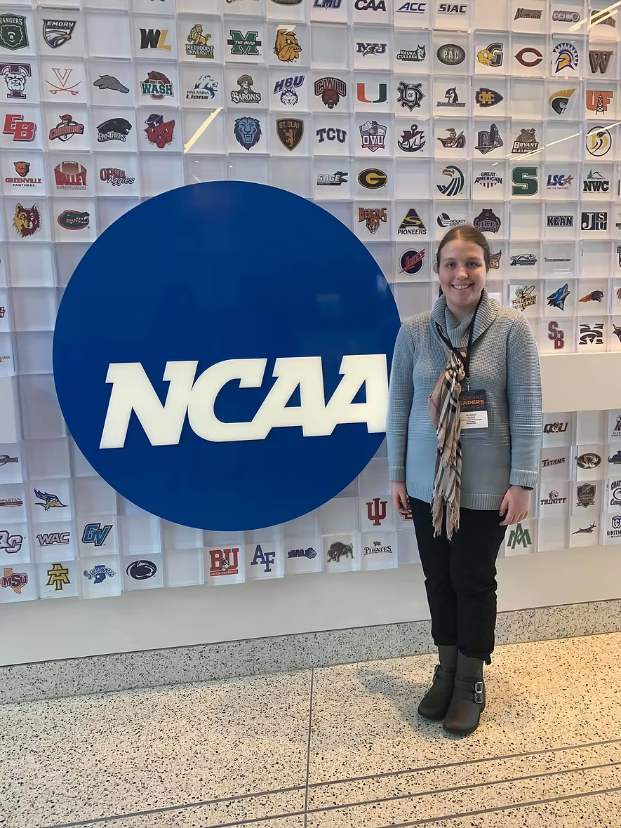 A young woman standing in front of a large NCAA logo and a display wall with college logos.