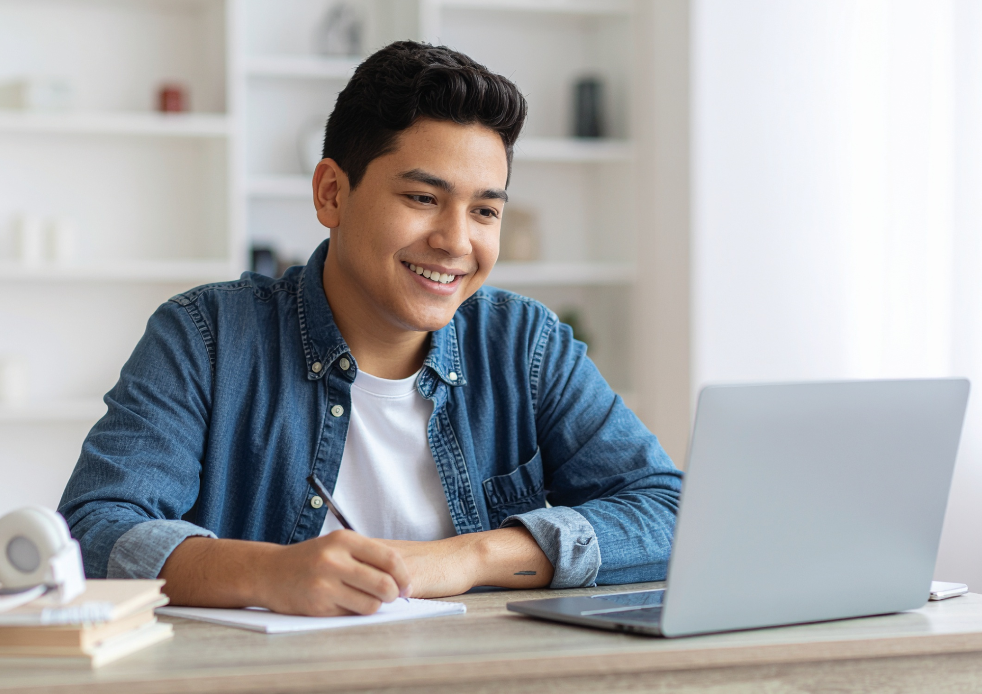 A young man with dark hair smiling at a laptop while writing on a notebook at a desk.