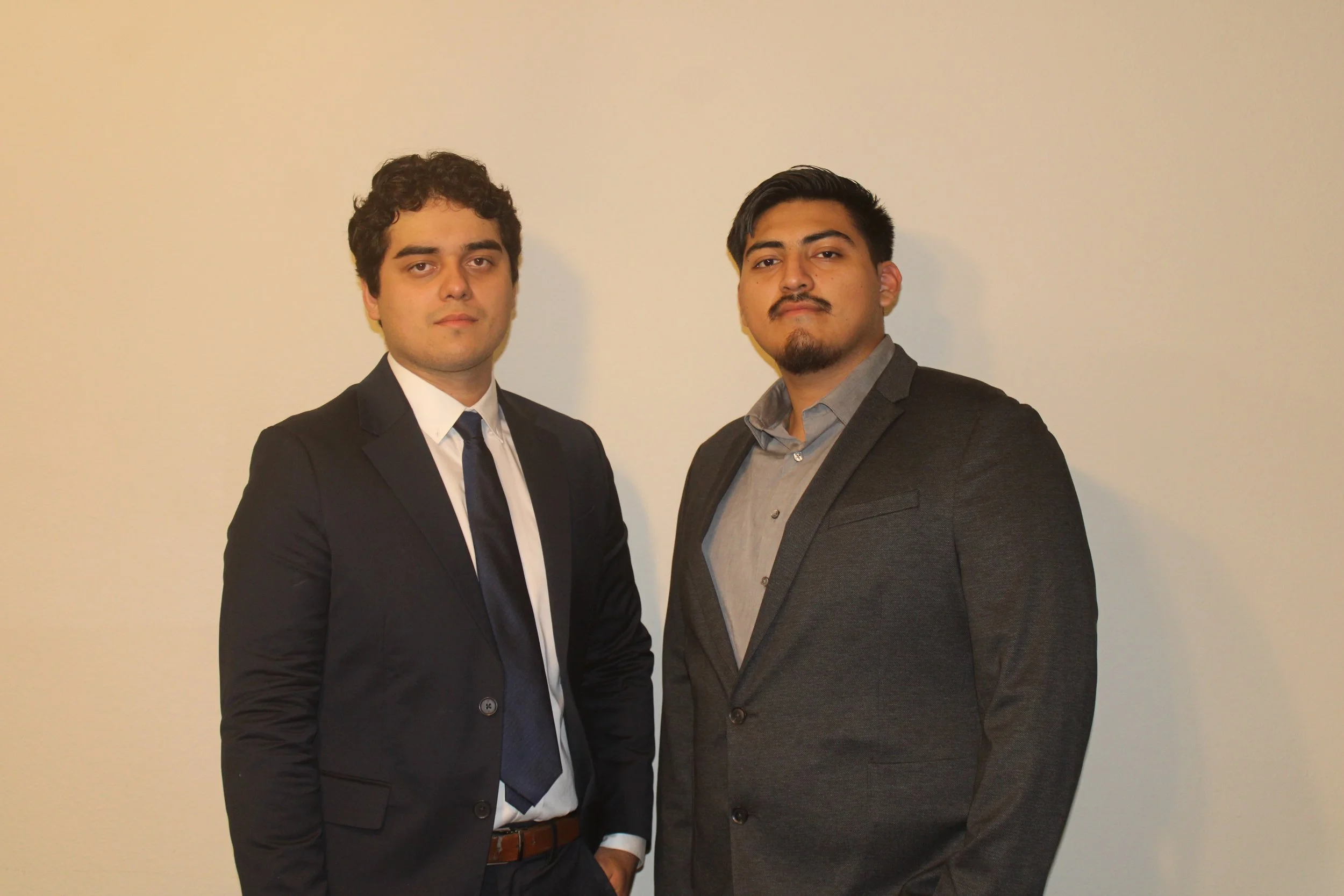 Two young men in formal suits standing side by side against a plain, light-colored wall, looking directly at the camera.