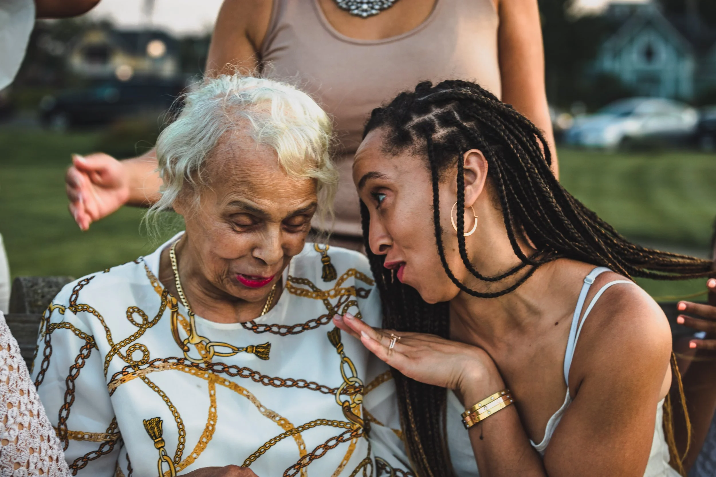 Two women leaning close together in a loving moment outdoors, one elderly with white hair, one younger with braided hair.