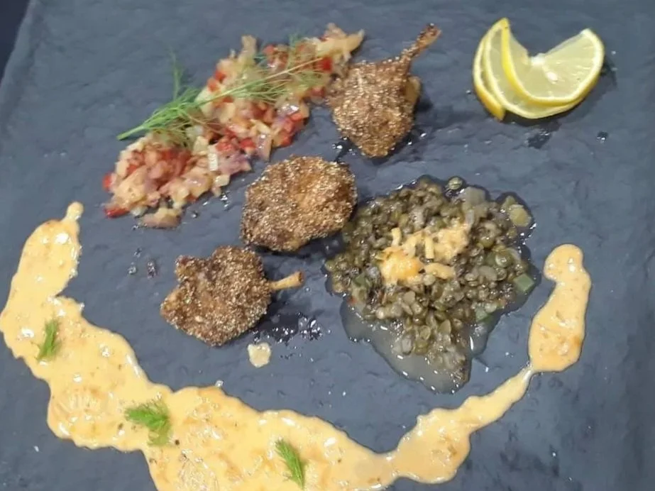 A gourmet dish on a black slate plate featuring three breaded and fried crab cakes, a lemon wedge, a colorful chopped tomato and onion relish topped with microgreens, a spoonful of creamy yellow sauce garnished with herbs, and a serving of seasoned lentils topped with melted cheese.