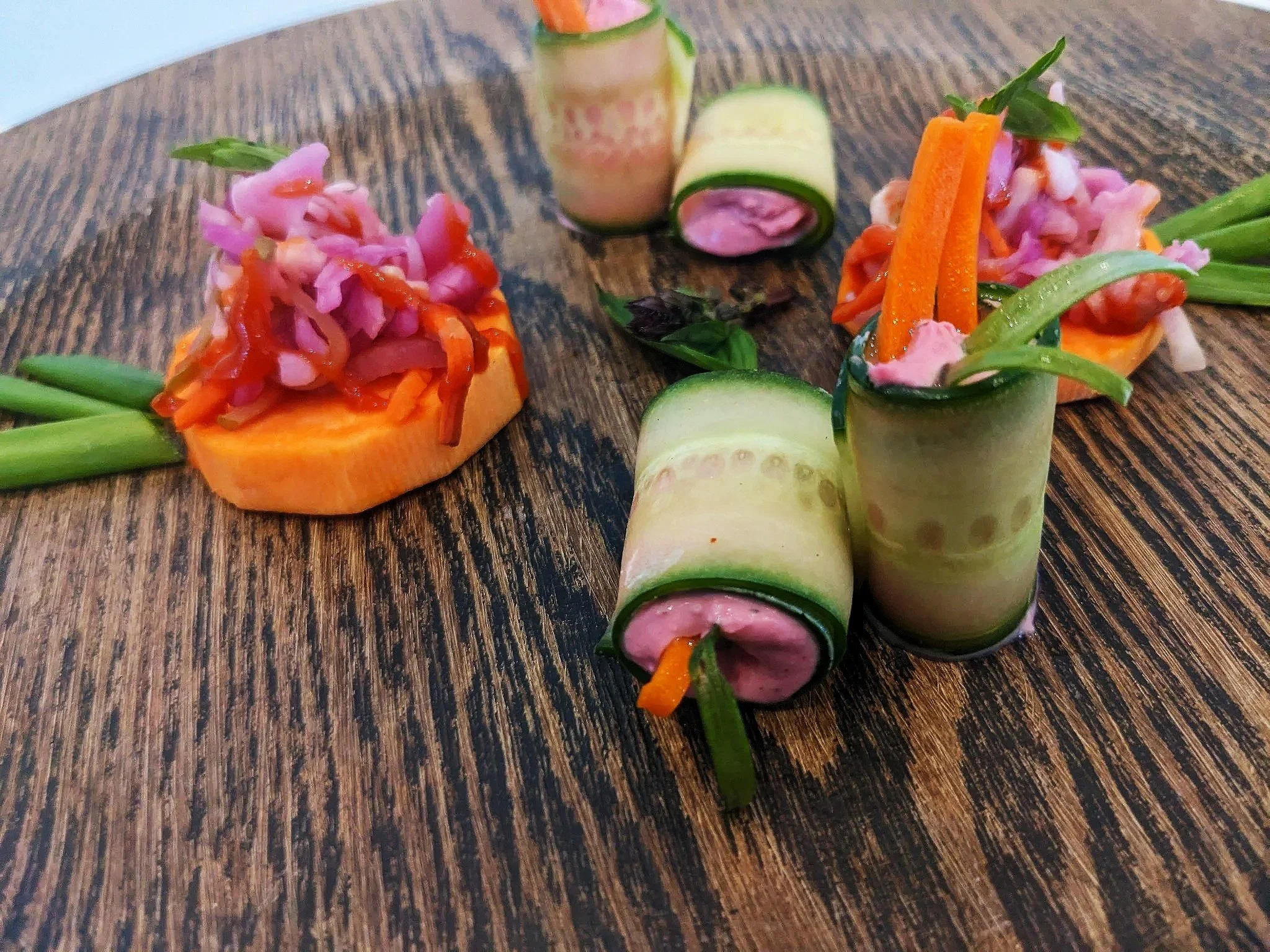 Vegetable tartlets topped with sliced pickles, carrots, pink pickled onions, and fresh herbs, arranged on a wooden surface.
