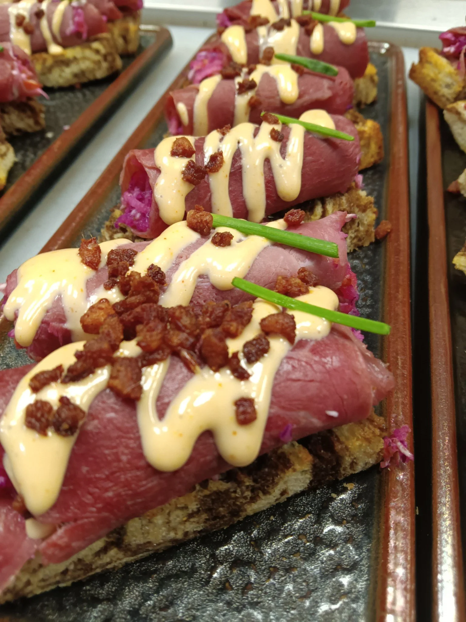 Close-up of slices of beef Wellington topped with a drizzle of creamy sauce, crumbled bacon, and garnished with a chive on a tray.