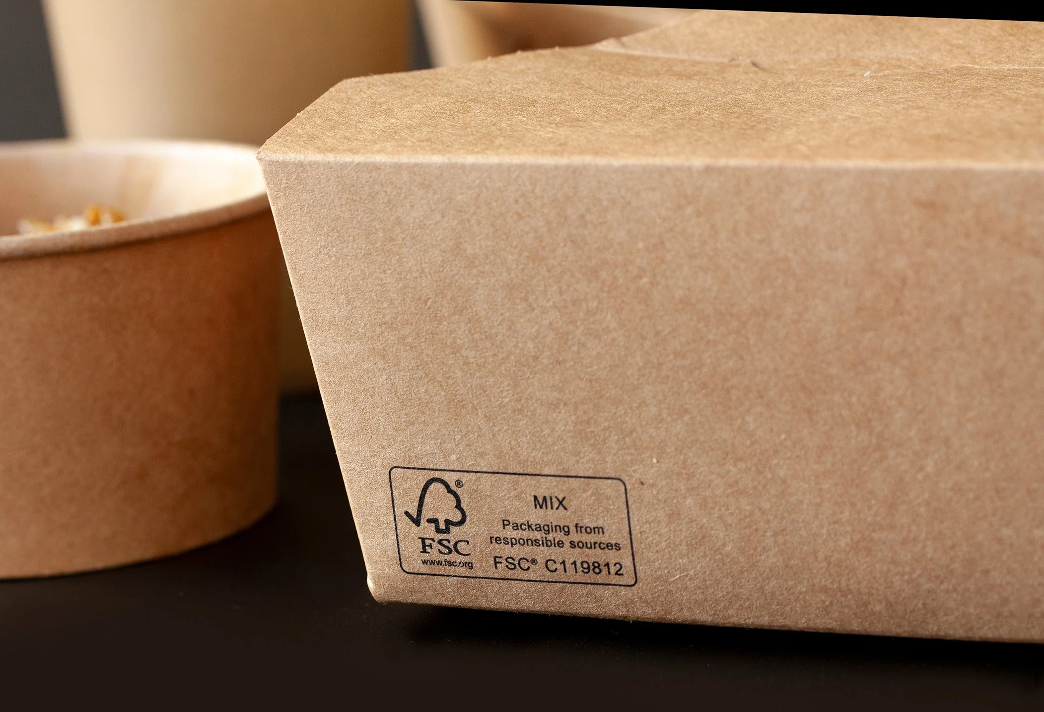 Close-up of a cardboard takeout container with a FSC certification label, partially visible, on a black surface.