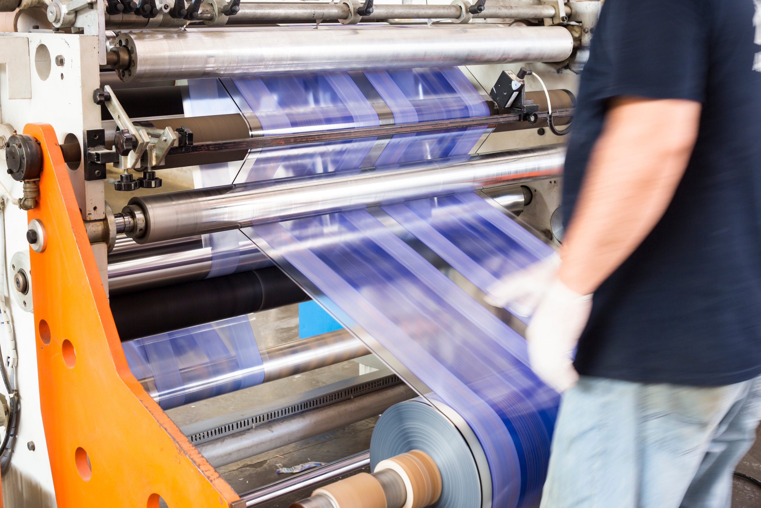 Industrial printing machine producing blue printed sheets or labels moving along rollers with a person inspecting or working on it.