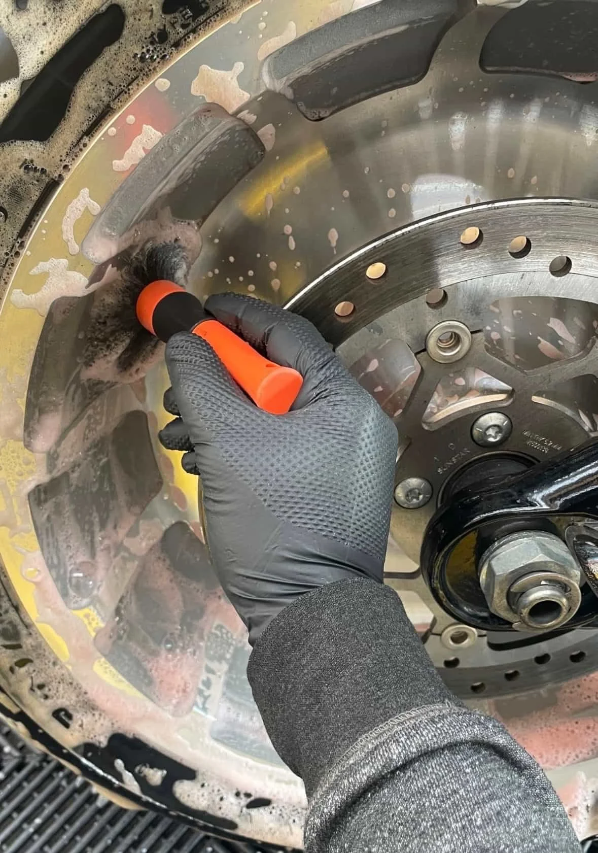 Une personne porte des gants noirs en nettoyant un disque de frein d'une moto avec une brosse de nettoyage orange.