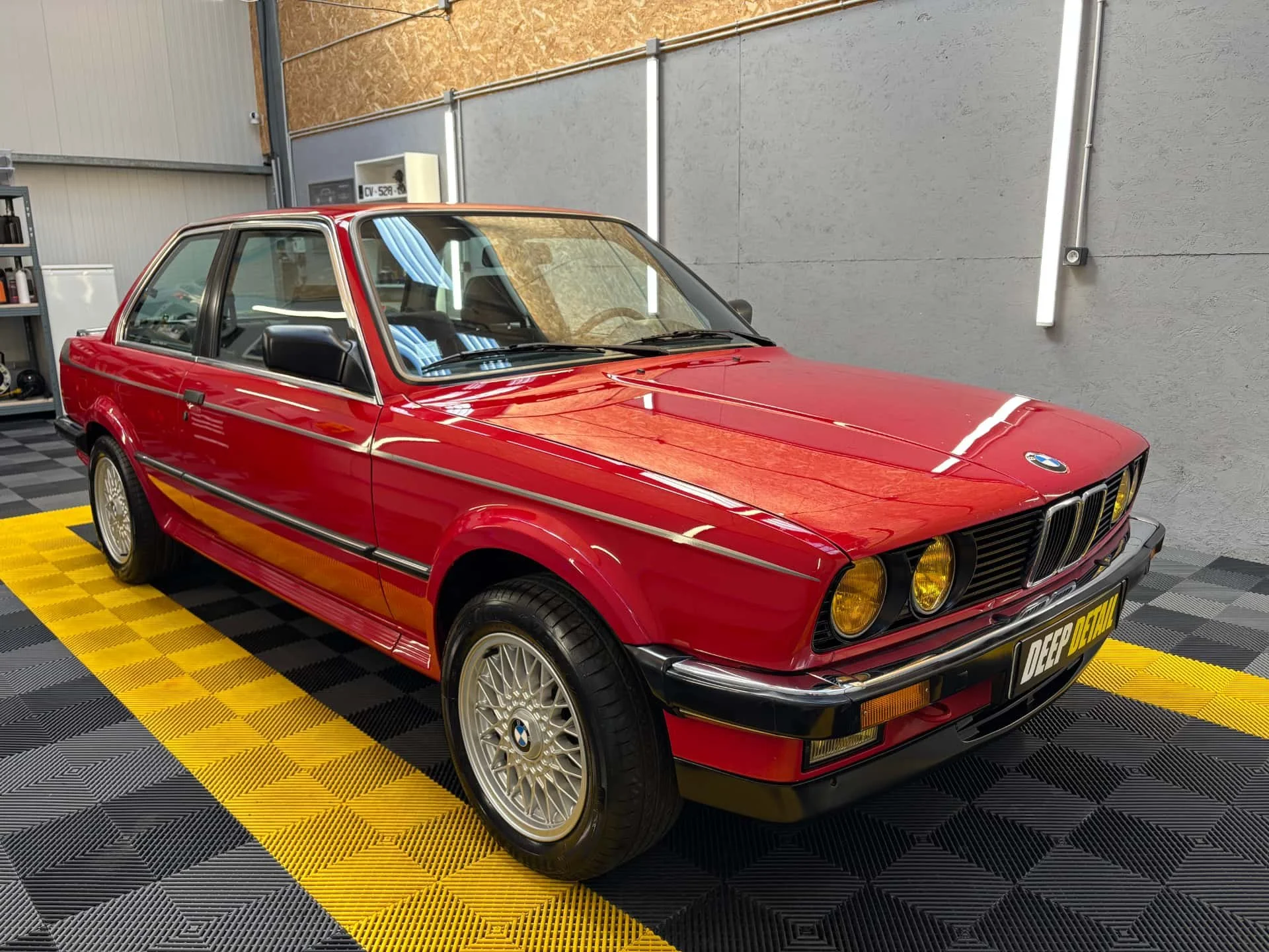 Une voiture BMW rouge ancienne dans un atelier, vue de face, sur un sol noir et jaune.