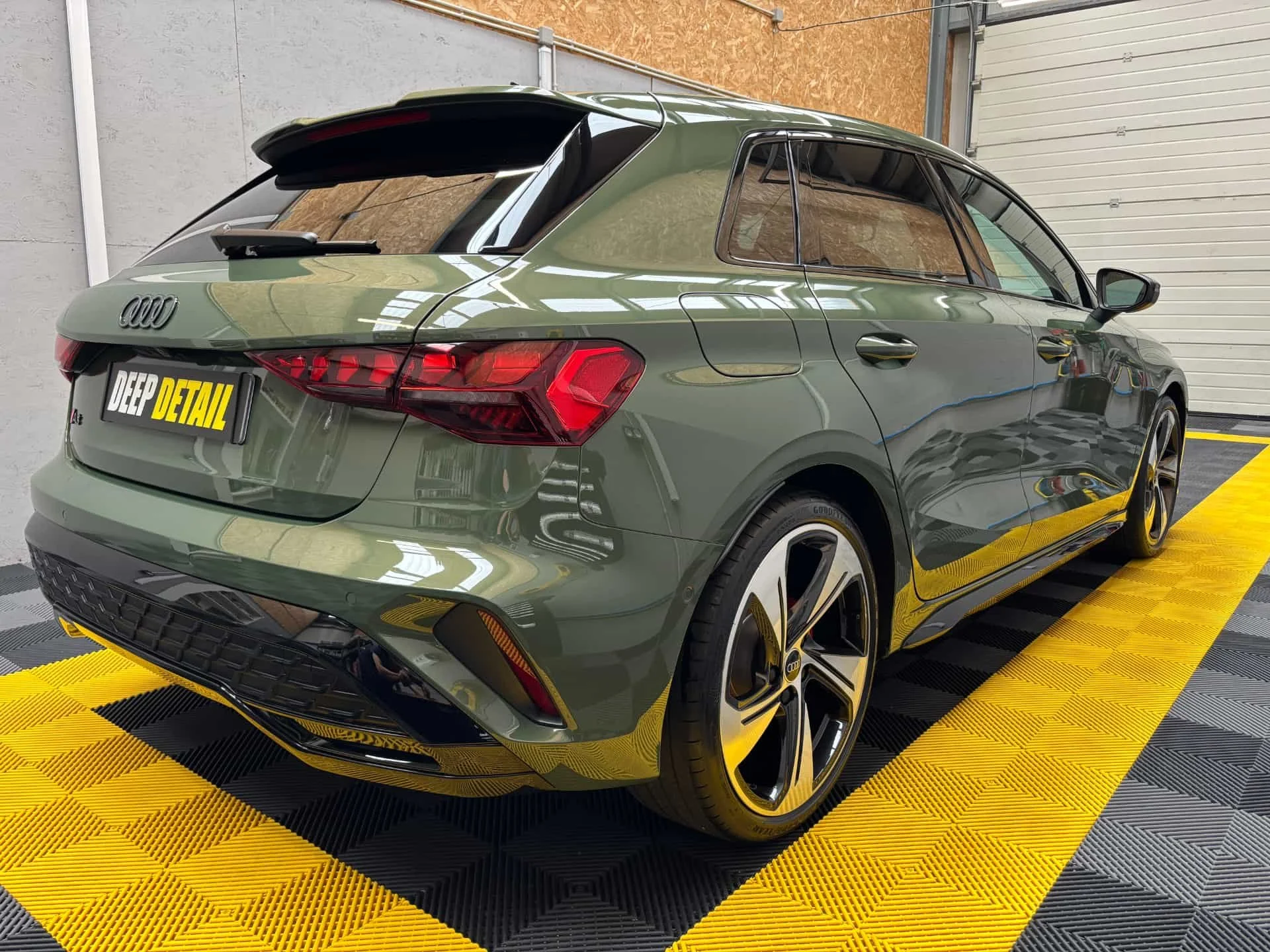 Voiture Audi verte en intérieur, stationnée sur un tapis noir et jaune, avec un mur en bois et un garage blanc en arrière-plan.