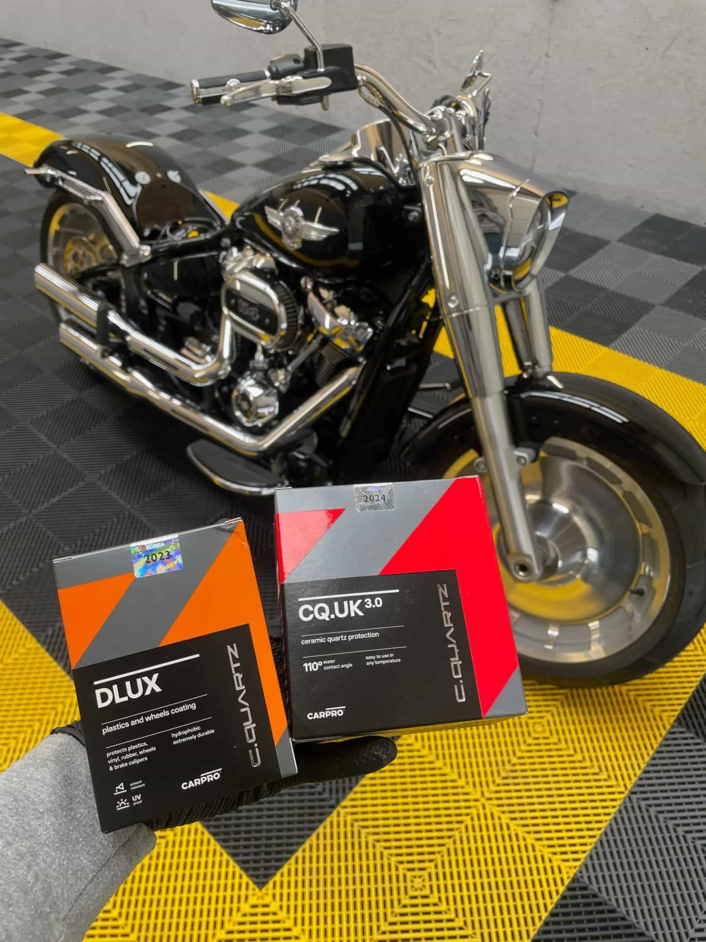 Une moto noire avec des éléments en chrome, placée sur un sol en avant-plan. Deux boîtes de produits CARPRO, une pour la protection des plastiques et moyeux avec une couleur orange, et une autre pour la protection en céramique du quartz avec une couleur rouge, sont tenues dans la main d'une personne, devant la moto.