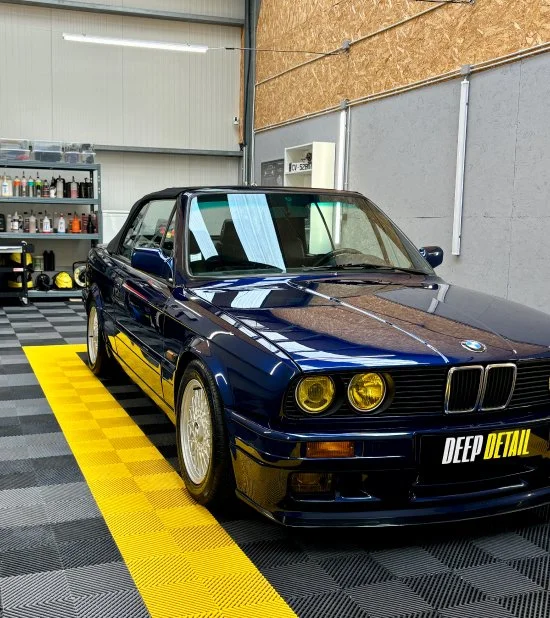 Voiture BMW classique bleu dans un atelier de nettoyage ou de detailing, avec un sol noir et jaune, étagères de produits de nettoyage en arrière-plan.