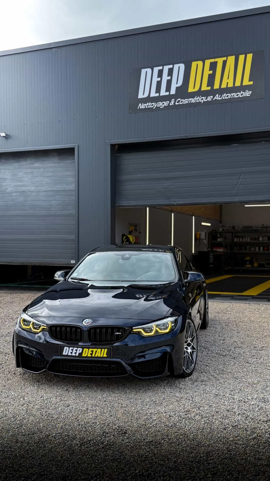 Une voiture noire BMW M4 devant un bâtiment gris avec un panneau de nettoyage automobile "DEEP DETAIL".