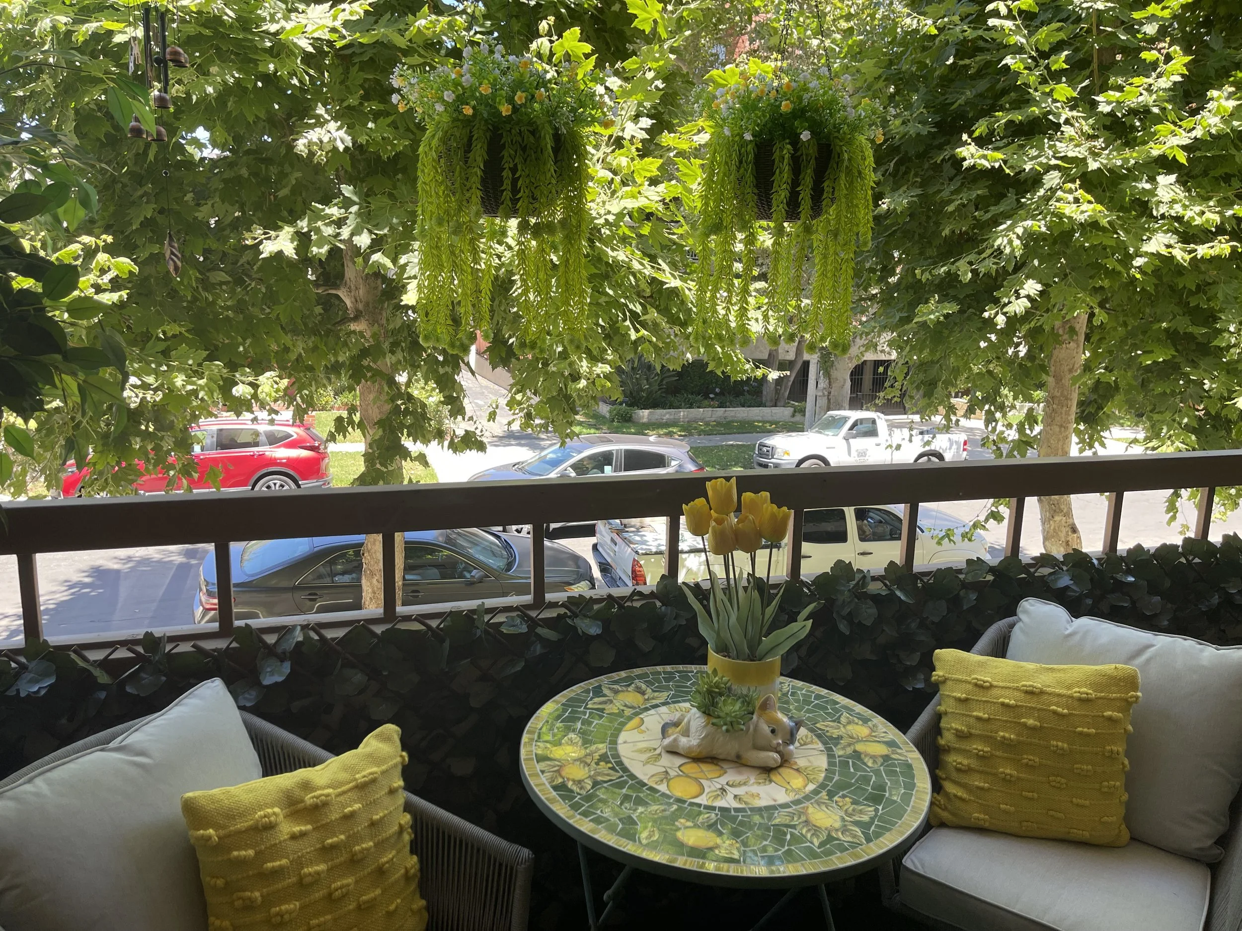 View of a balcony with a round table with a colorful lemon-themed mosaic top, surrounded by cushioned chairs with yellow pillows, overlooking a street with parked cars and lush, green trees. Hanging flower baskets with green foliage and white flowers