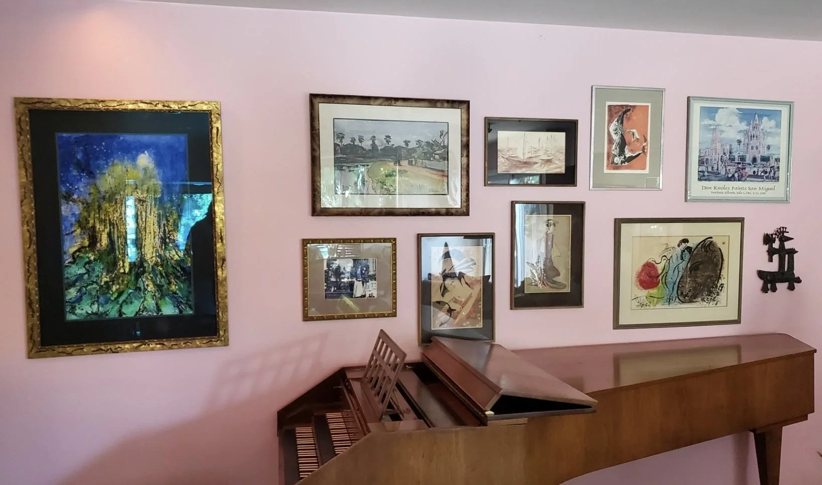 A pink wall decorated with various framed artworks, including paintings and illustrations, and a small wooden piano against the wall.