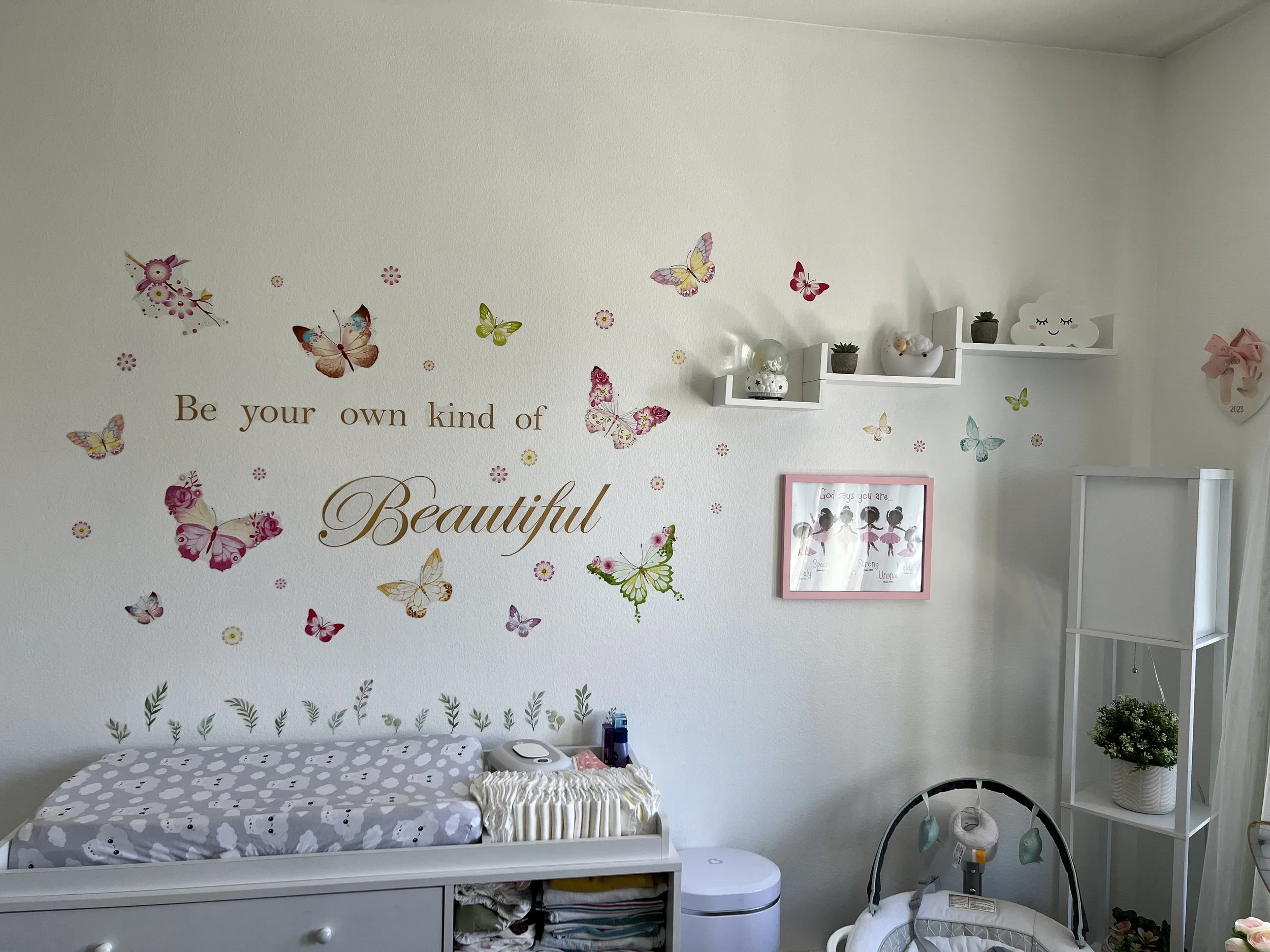 A nursery room with a white changing table with a gray polka dot pad, a white shelf with potted plants, a cloud-shaped decor, and butterfly stickers on the wall with a partial view of a framed picture and a white shelf.