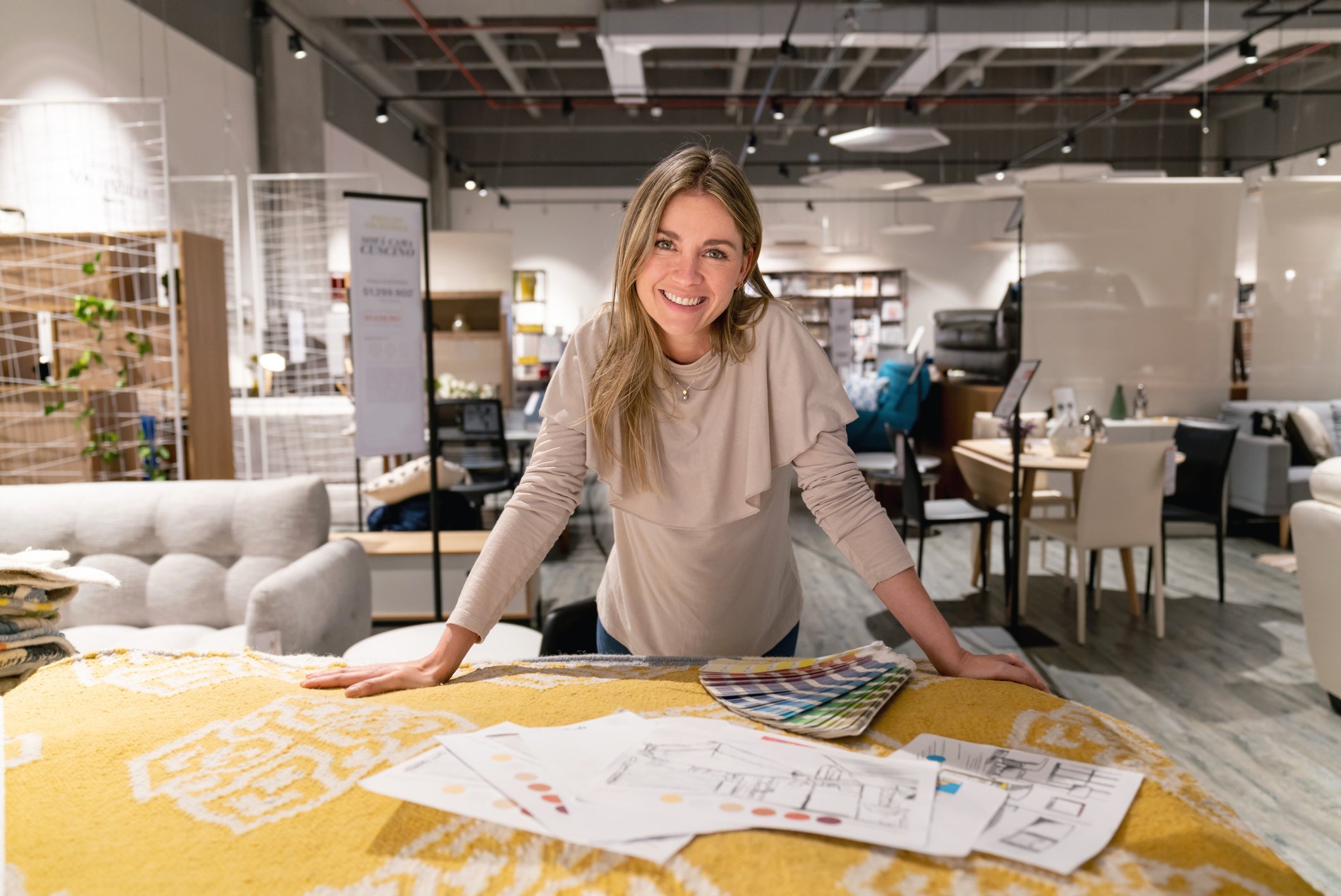 A woman with light brown hair smiling and leaning on a table in a furniture store, surrounded by fabric samples and design plans.