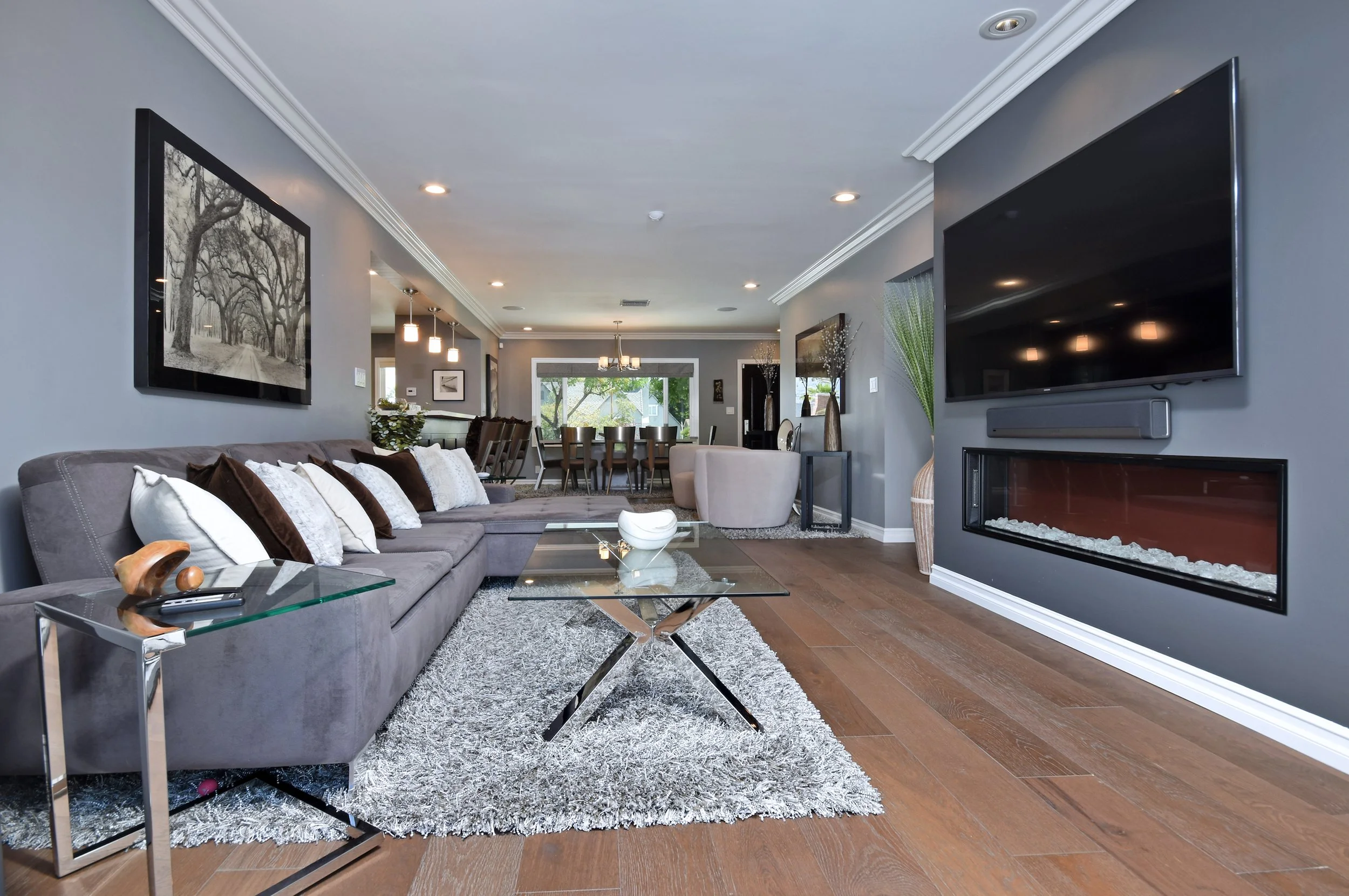 Living room with gray sofa, glass coffee table, large wall-mounted TV, fireplace, paintings, and dining area with windows and outdoor greenery visible