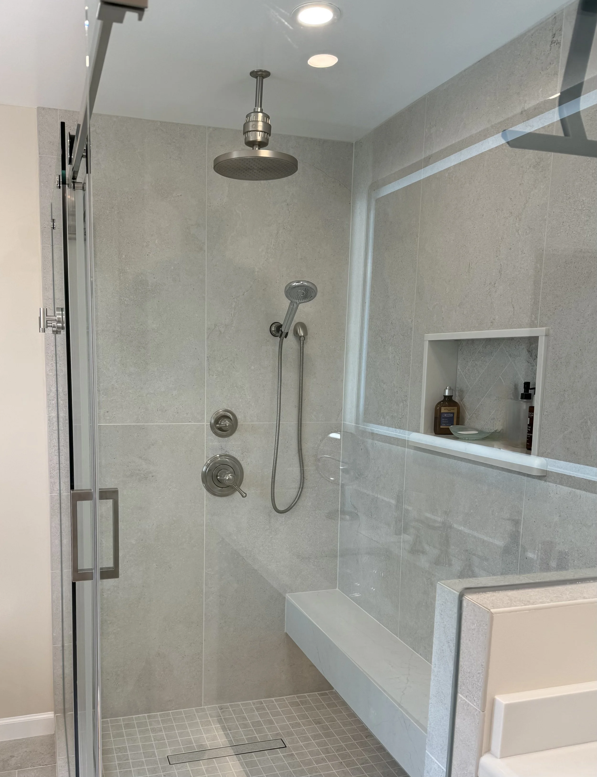 A modern walk-in shower with a glass door, a large rainfall showerhead, a handheld showerhead, and a small recessed shelf with toiletries, all surrounded by light gray tiles.