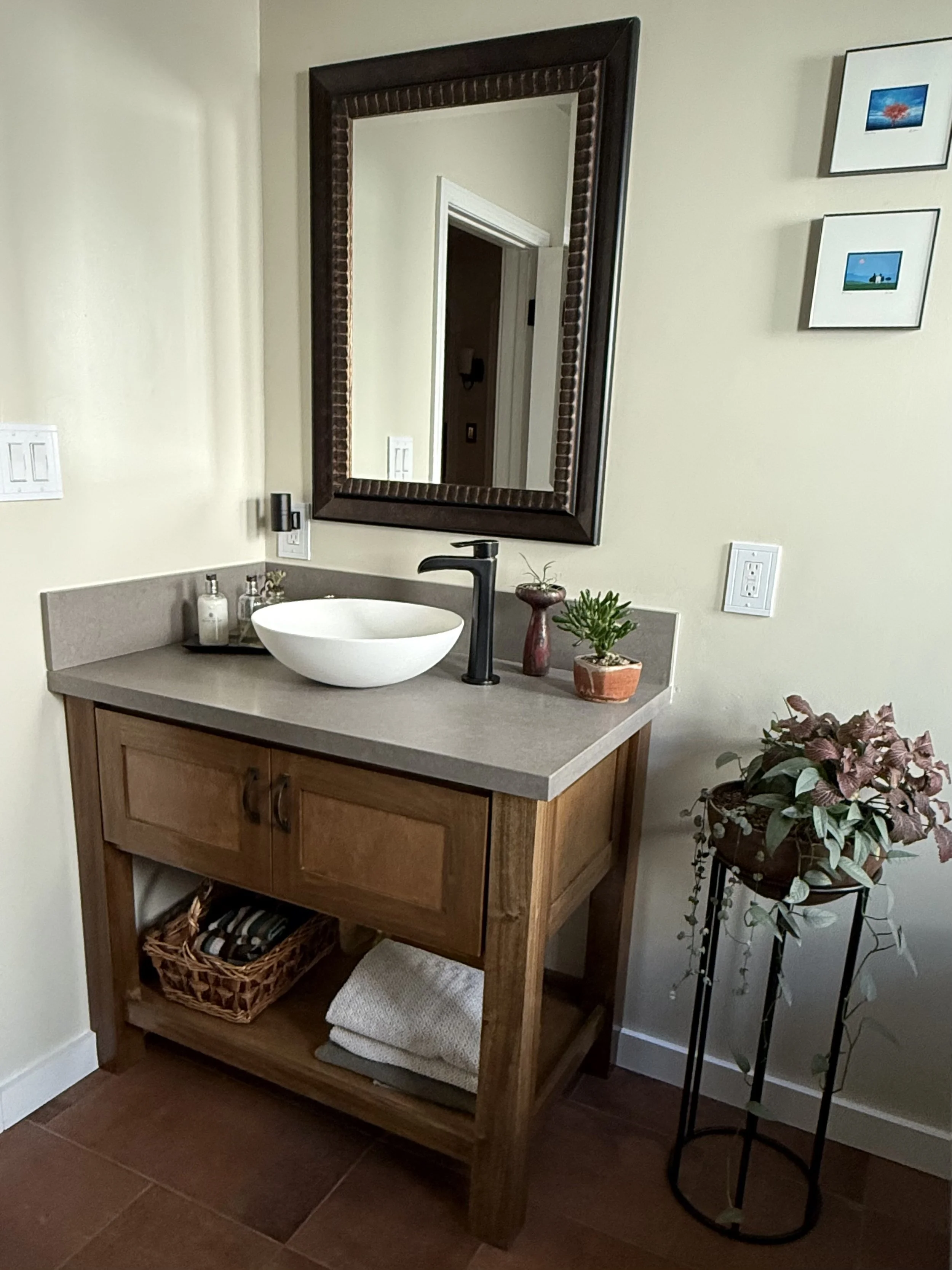 Bathroom vanity with a wooden cabinet, white Vessel sink, black faucet, mirror with dark wooden frame, small plants, soap pump, and towels underneath, next to a decorative plant stand with leaves.