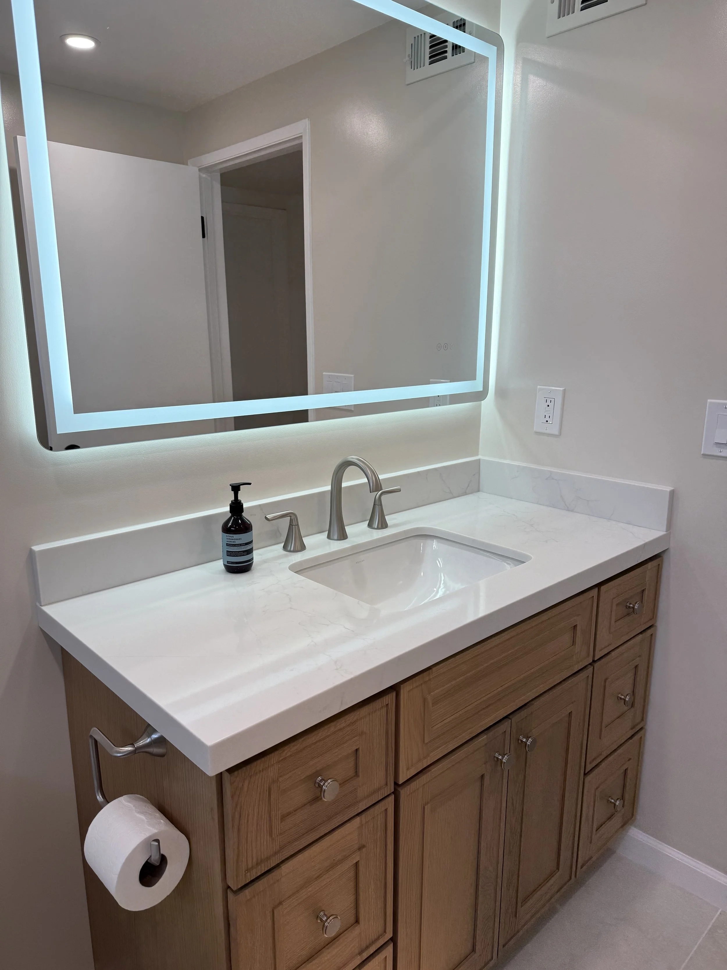Bathroom vanity with a wooden cabinet, white marble countertop, a sink, a modern silver faucet, a mirrored illuminated medicine cabinet, a soap dispenser, a wall-mounted toilet paper holder with a roll of toilet paper, and electrical outlets.